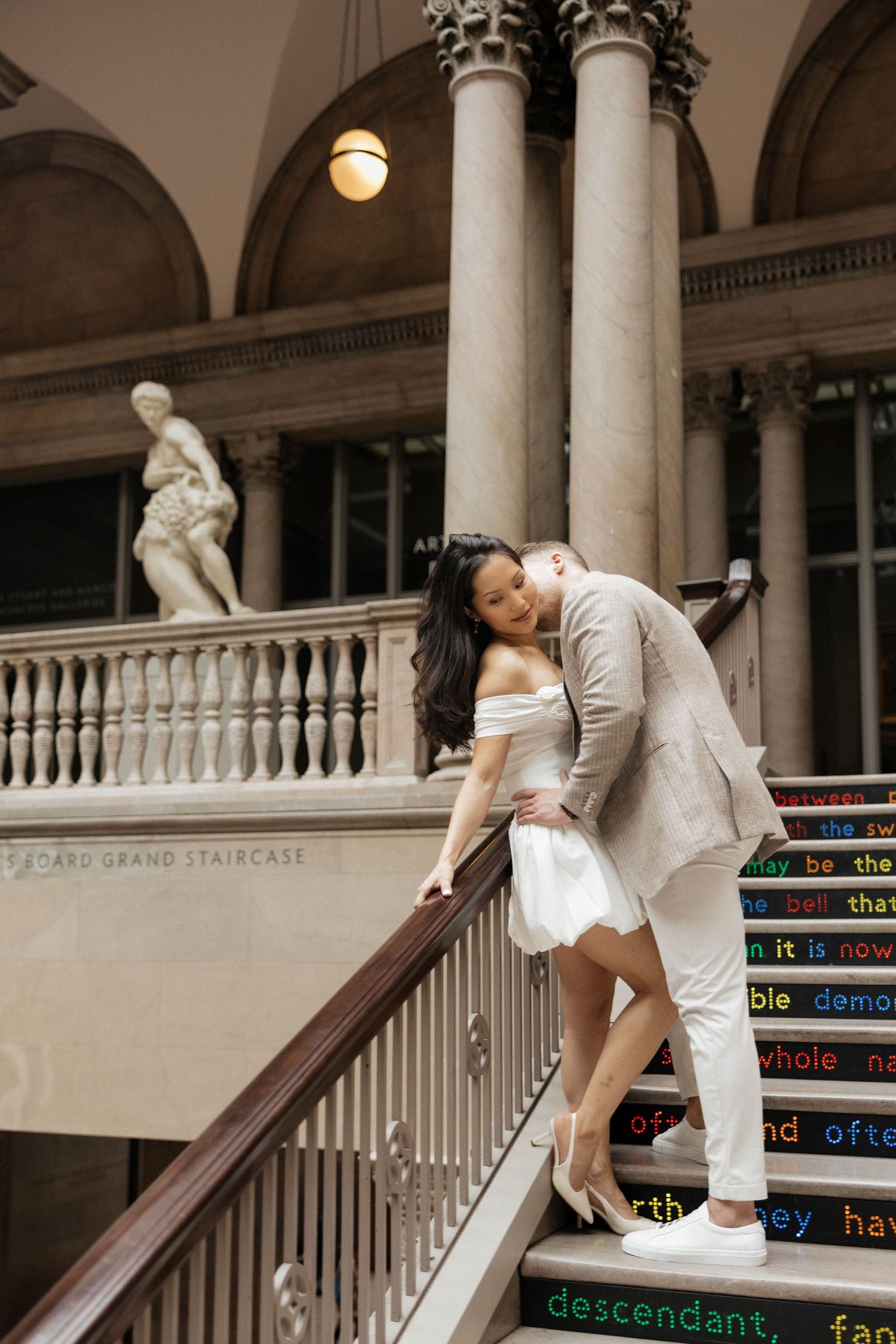 Romantic couple embracing on ornate museum staircase with classical columns and sculpture in background.