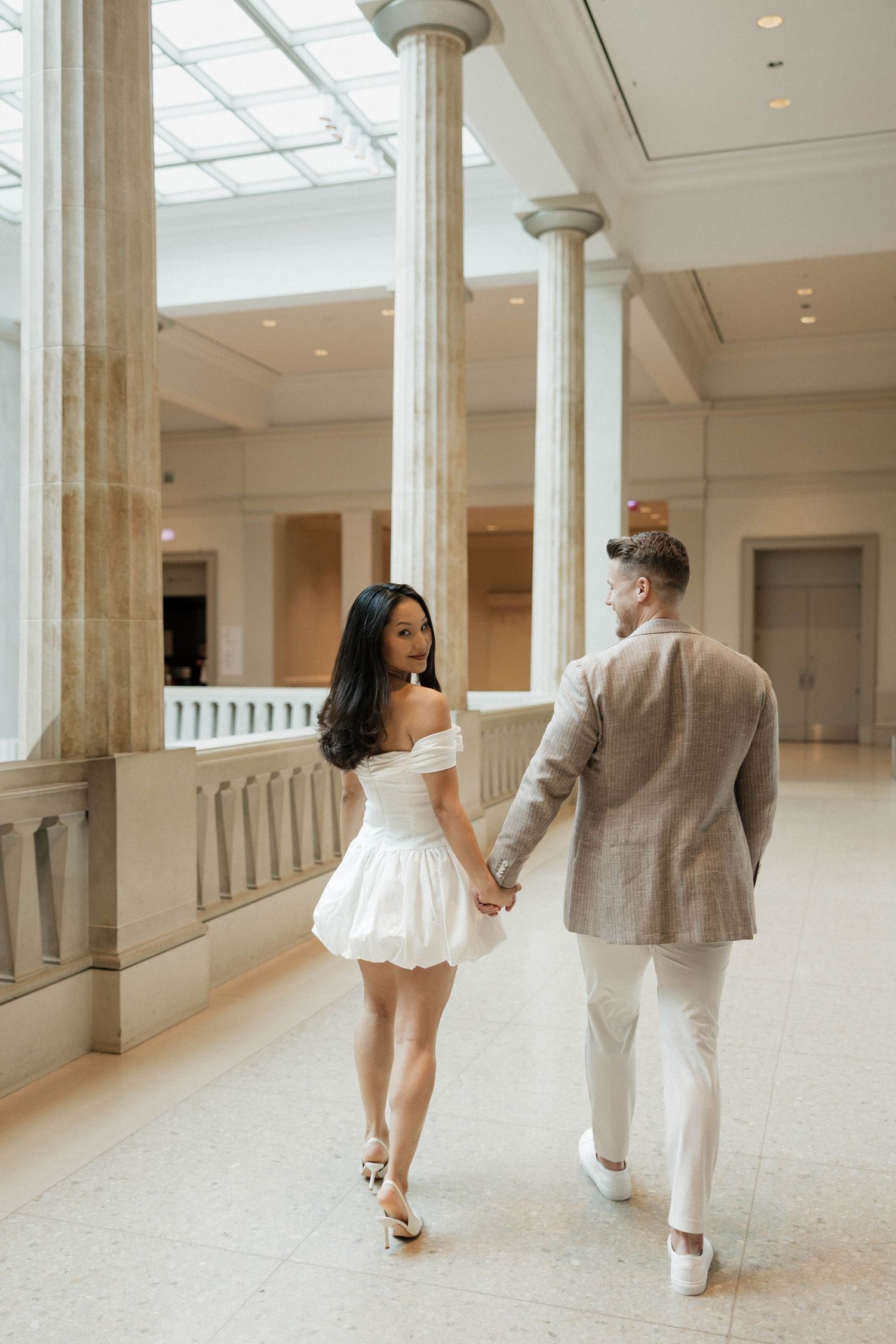 Couple holding hands walking through elegant hall with tall columns, woman in white mini dress, man in beige suit.