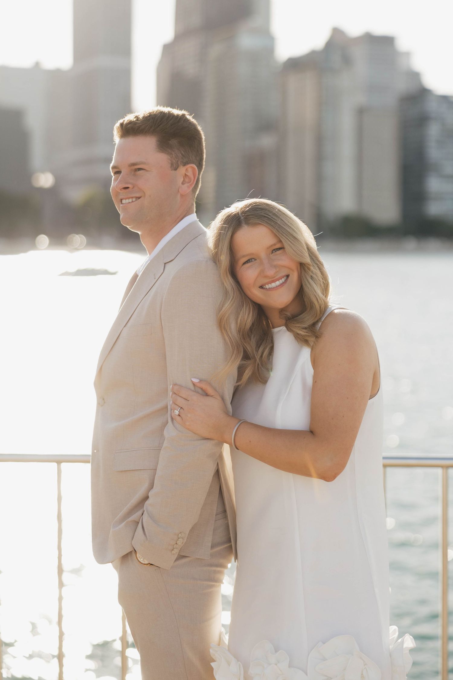 Couple posing by waterfront with city skyline, woman in white dress hugging man in tan suit, smiling outdoors.