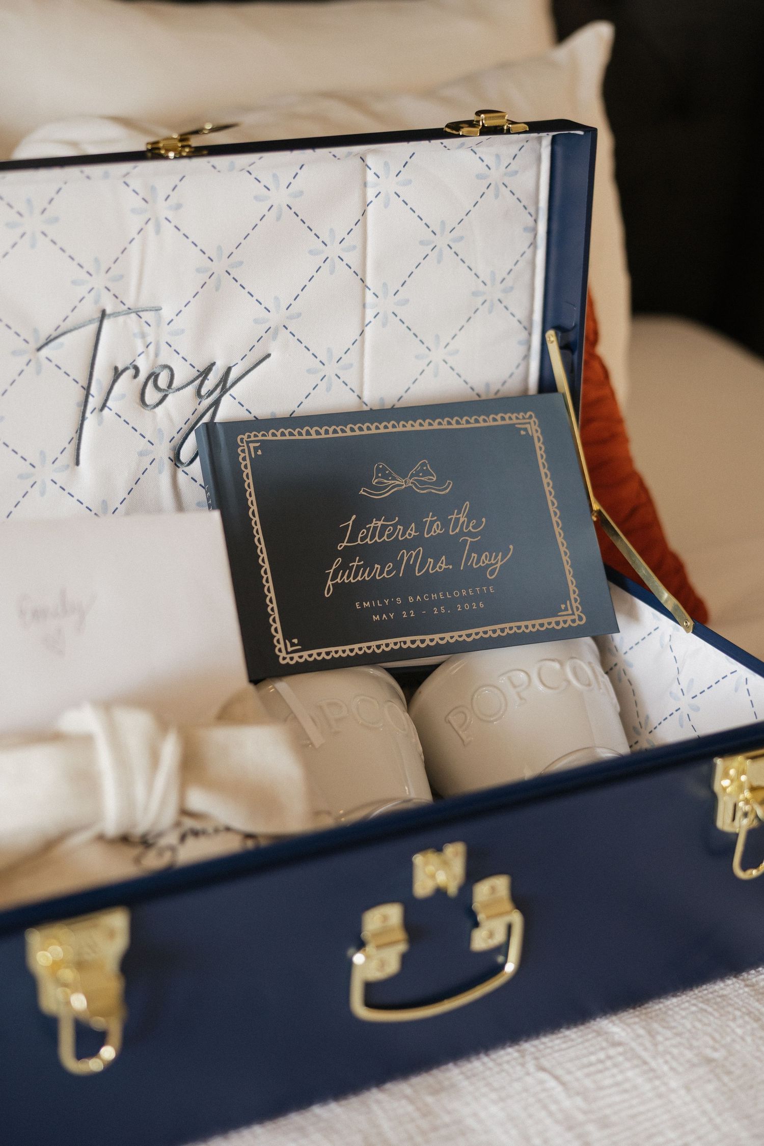 Navy blue suitcase with gold hardware containing personalized wedding gifts, monogrammed pillow, and Letters to the Future Mrs. Troy card.