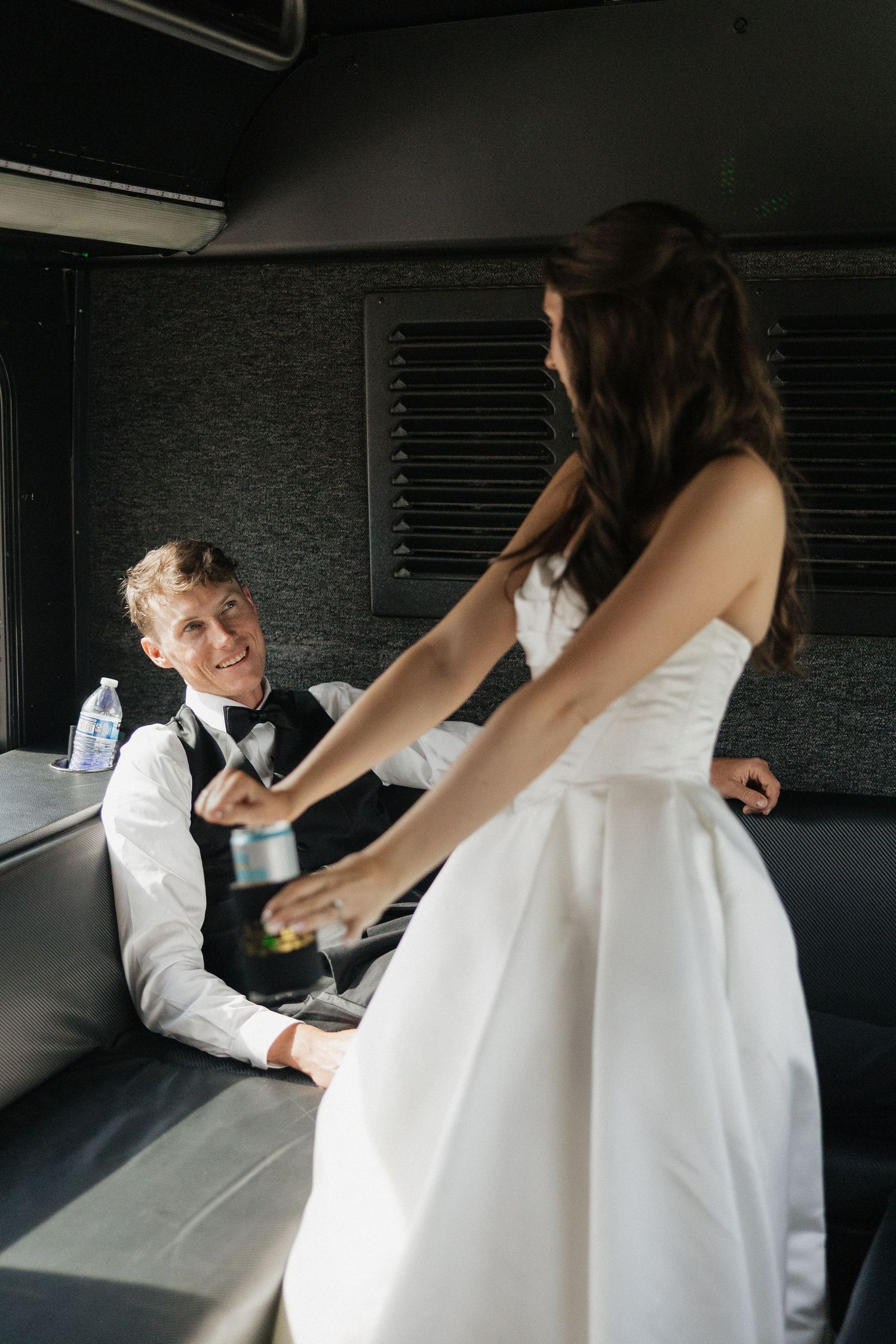 Bride in white gown and groom in vest share a drink inside a limousine on their wedding day.