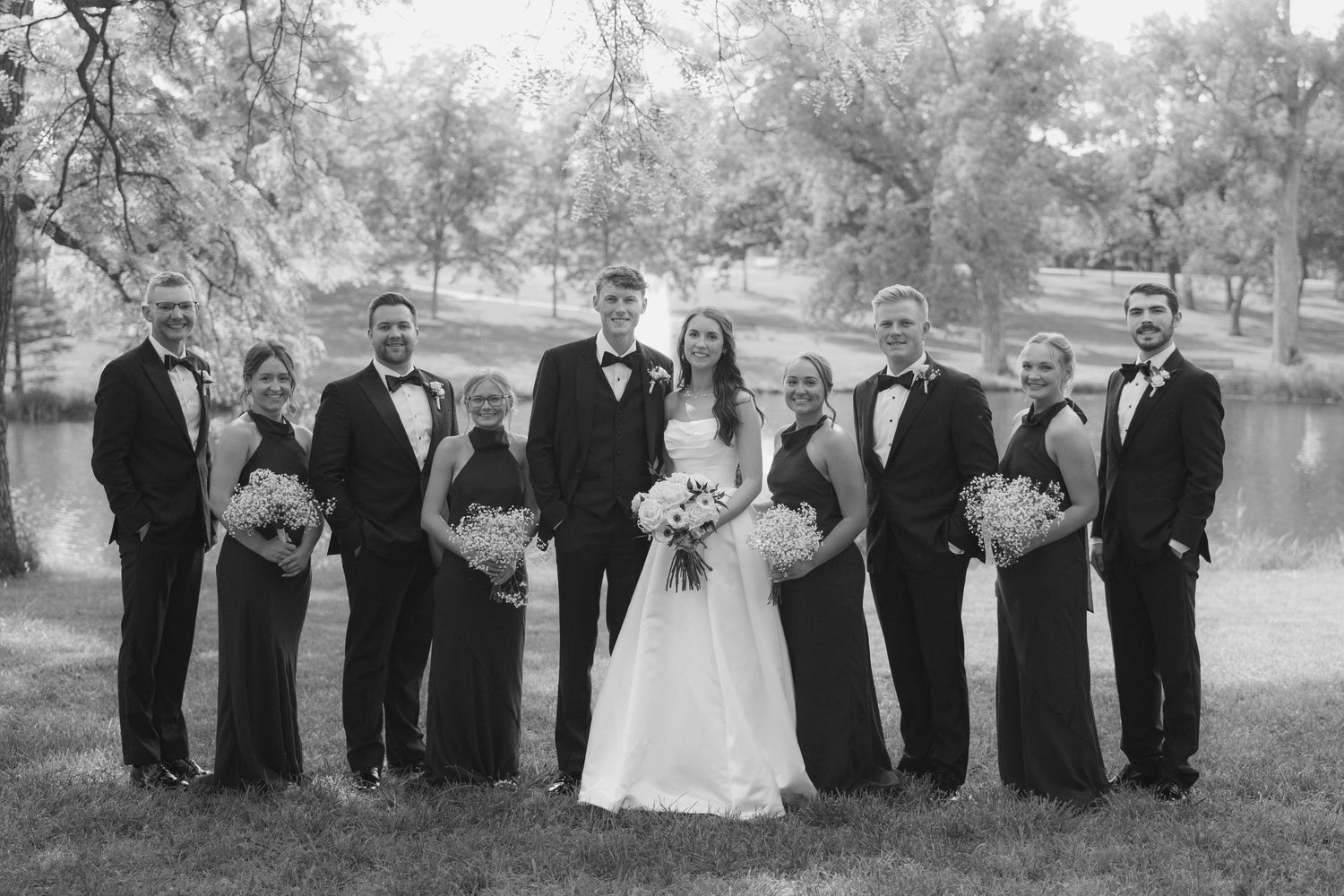 Black and white wedding party photo with bride, groom, bridesmaids in dark gowns and groomsmen in tuxedos outdoors.