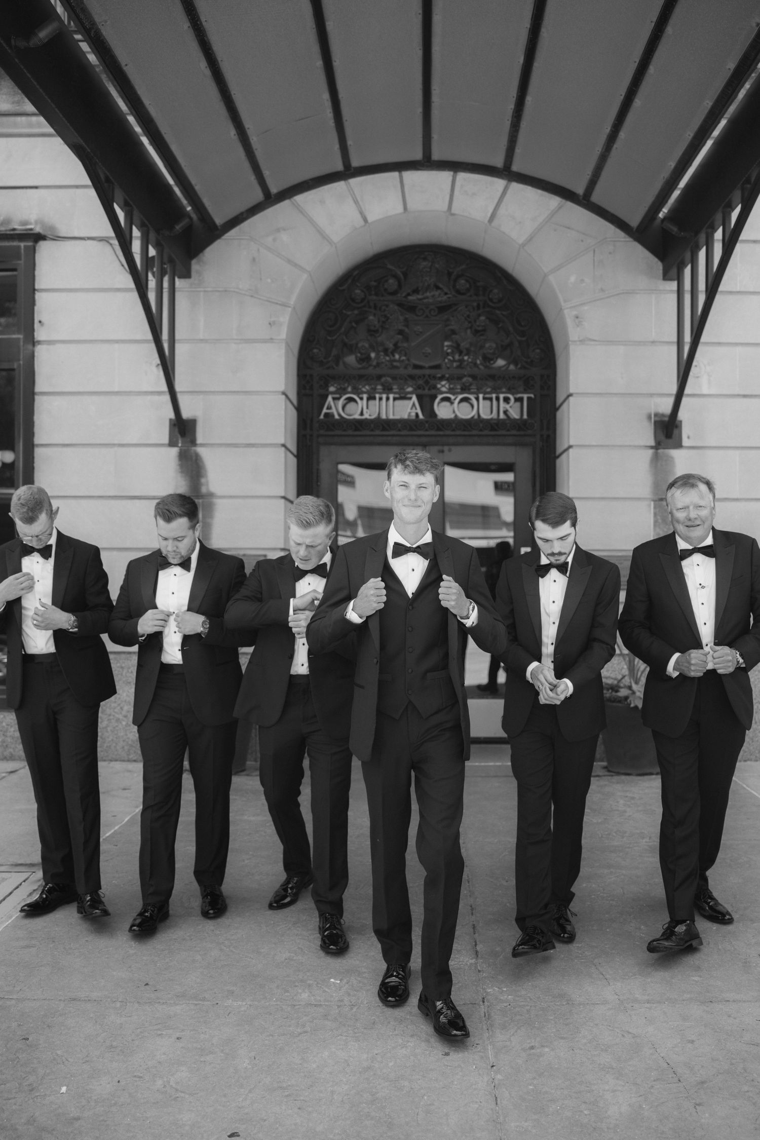 Groom and groomsmen in black tuxedos pose outside Aquila Court in a stylish black and white wedding photo.