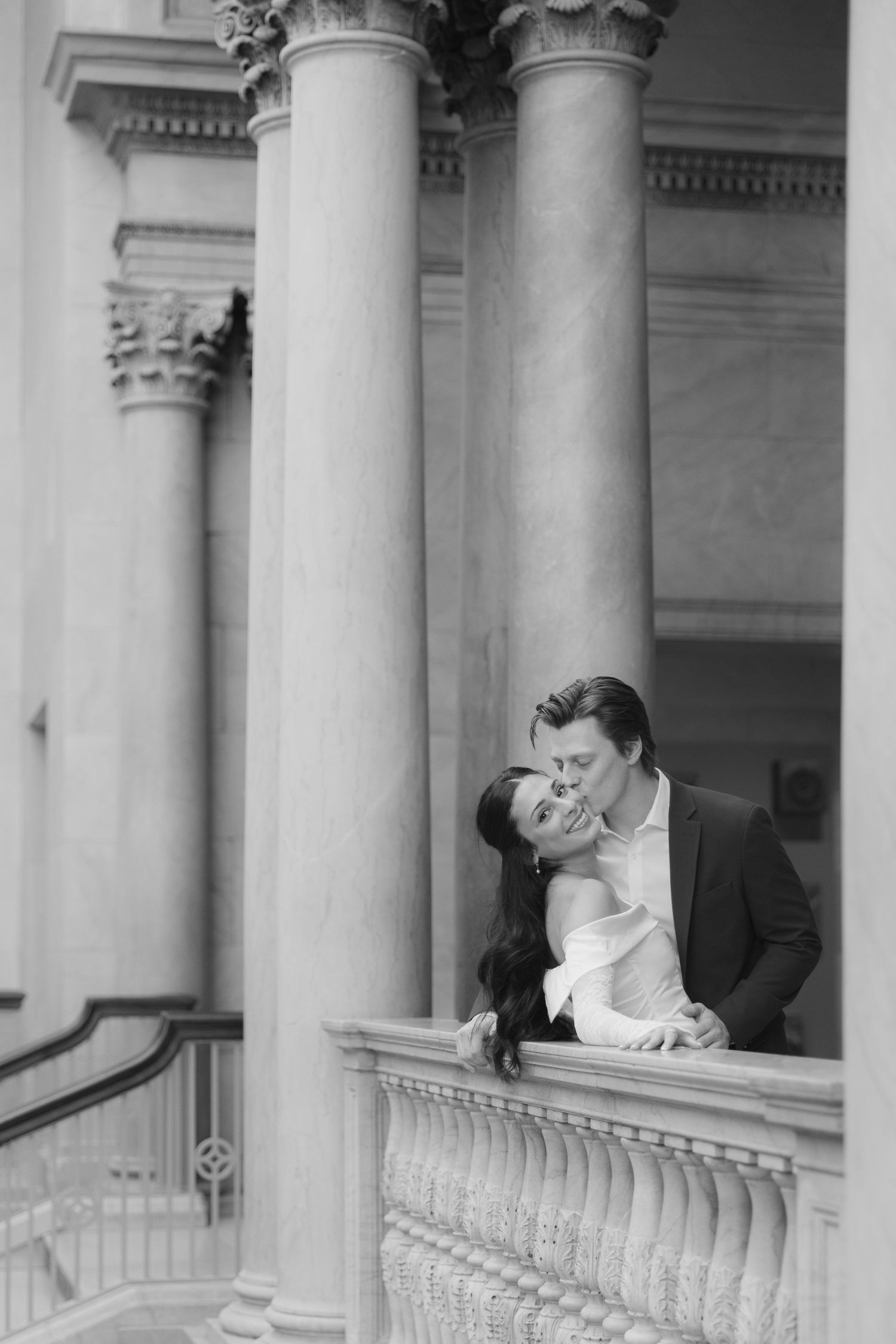 A romantic couple shares an intimate moment on a grand balcony with classical columns in black and white photography.