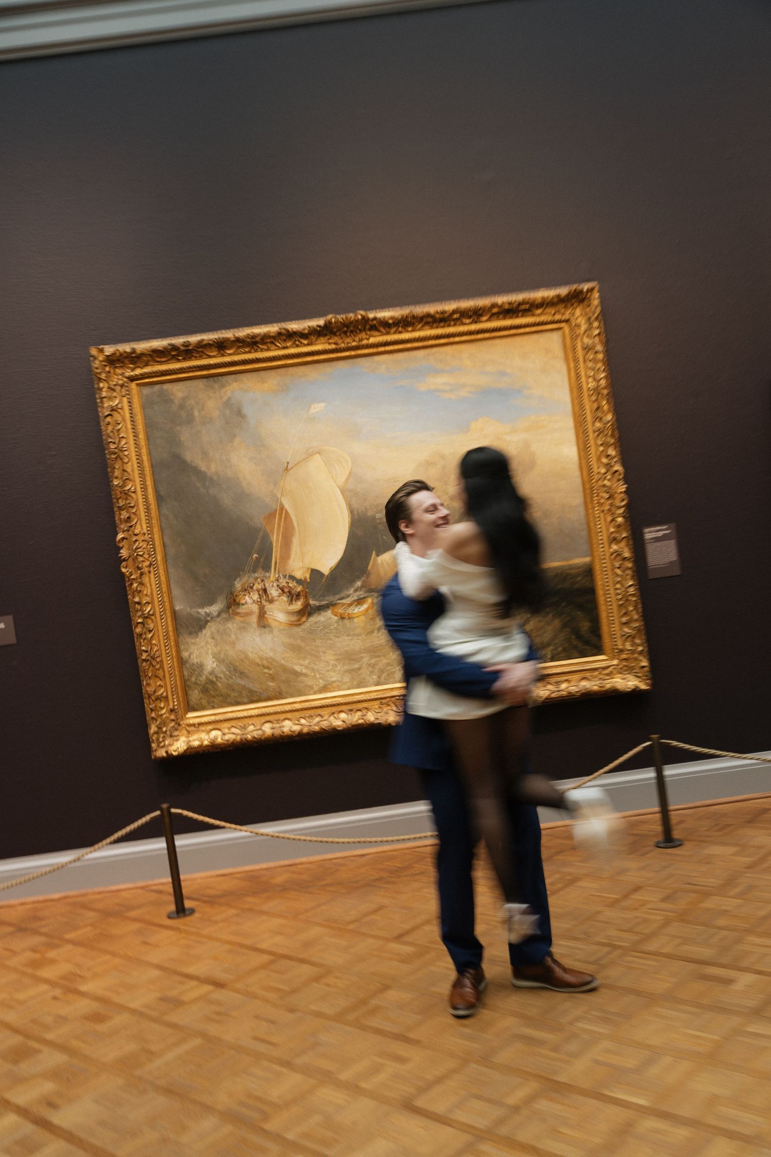 Blurred figures dance in front of large framed paintings in an art gallery.