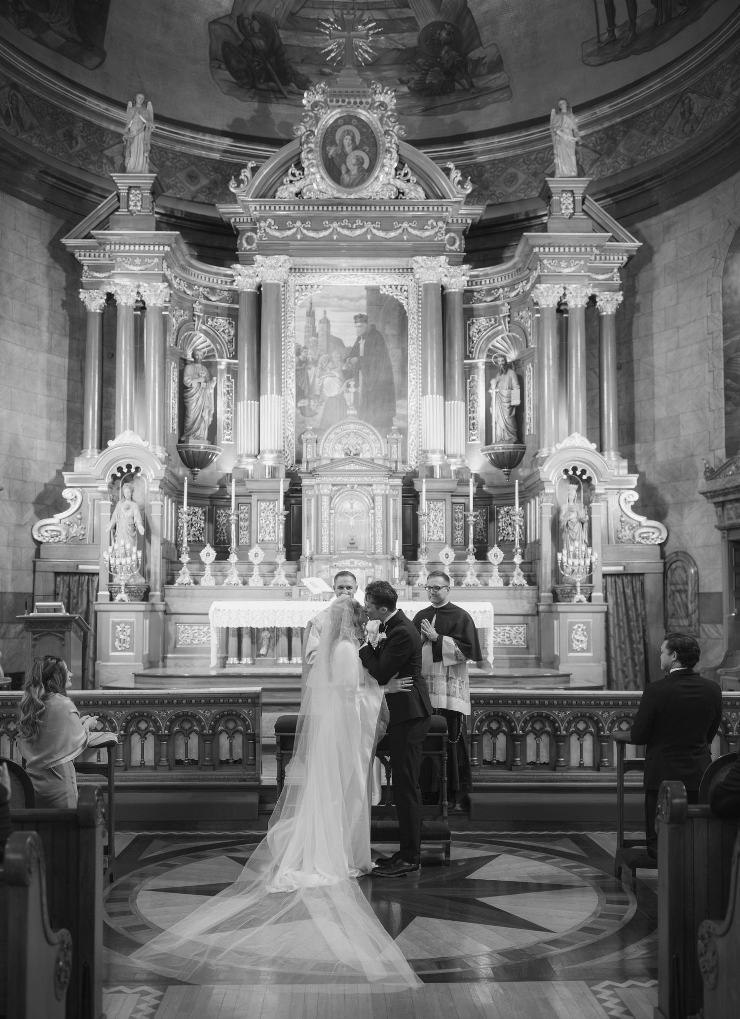 A black and white photograph of a wedding ceremony inside an ornate Catholic church with a grand baroque altar.