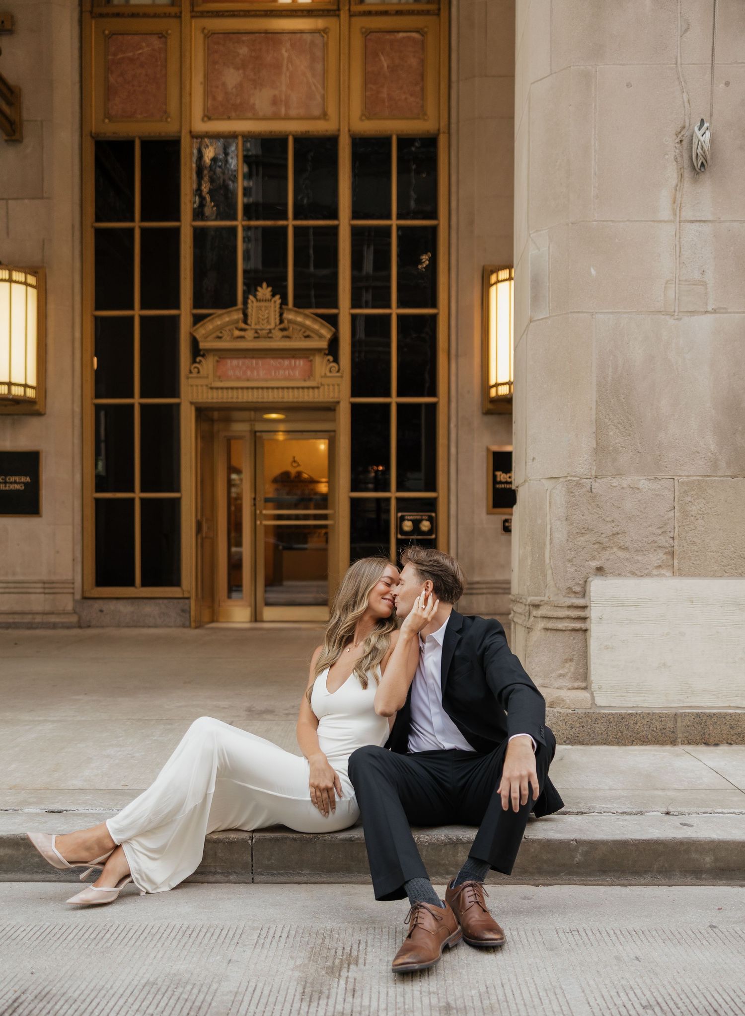 Couple in white jumpsuit and dark suit sit close on a stone step outside an ornate historic building entrance for an engagement photo.