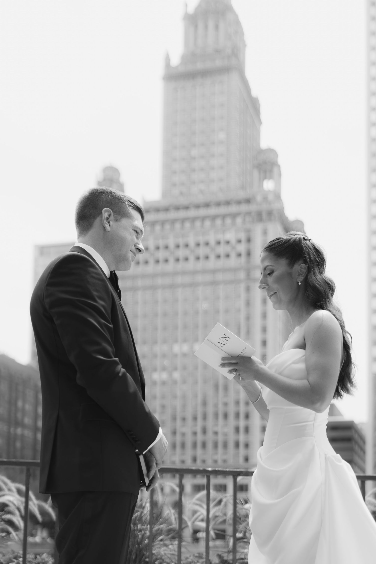 Black and white photo of a bride in a strapless gown reading her vows to a groom in a suit on a city terrace with a skyscraper behind.