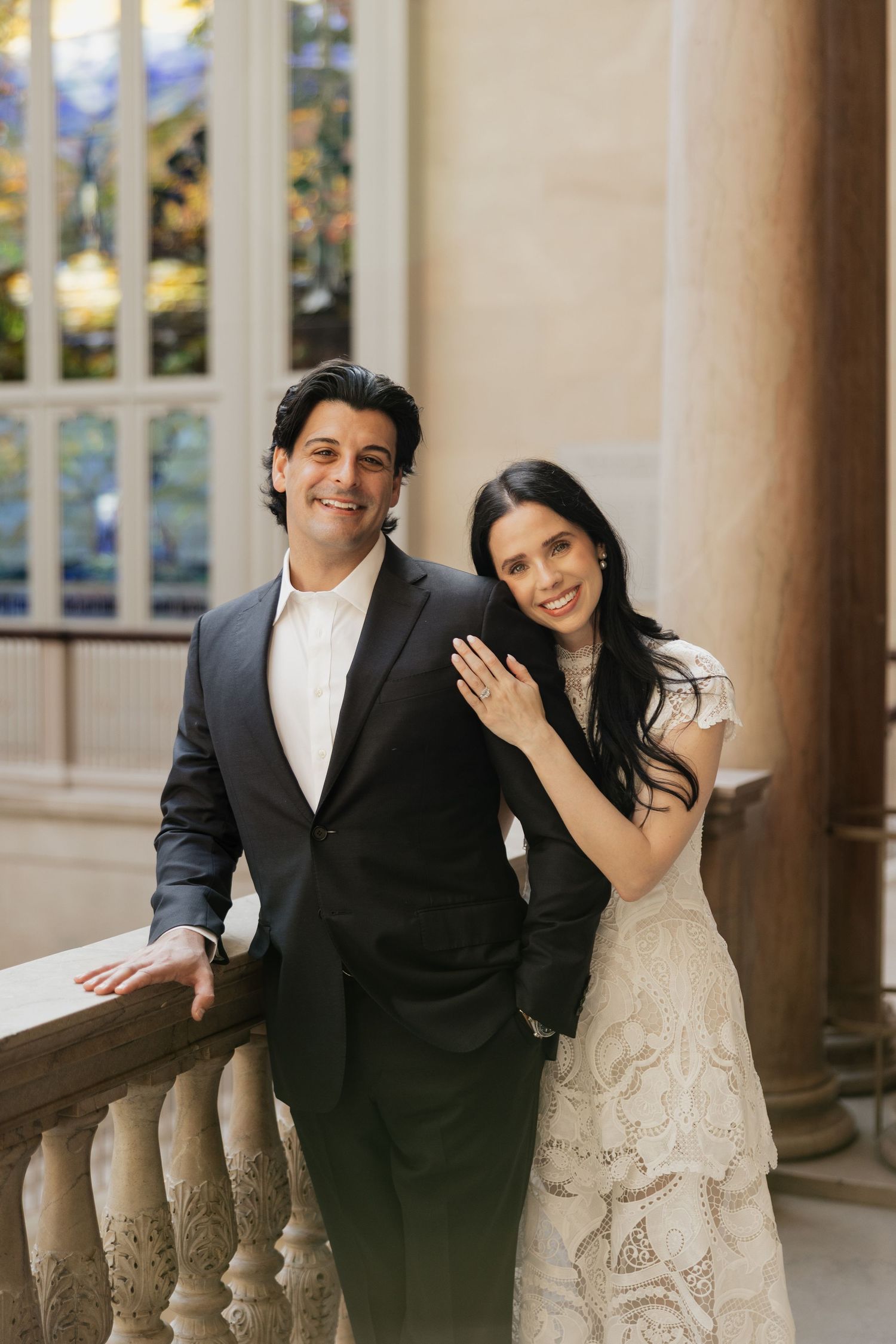 Smiling couple posing on a stone balcony, the man in a dark suit and the woman in a white lace dress leaning on his shoulder.