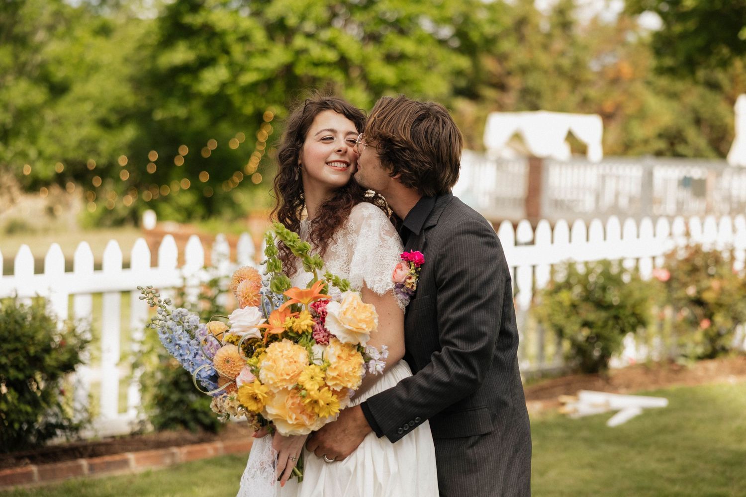 A groom kisses his smiling bride holding a colorful bouquet of yellow and pink flowers in a garden wedding setting.