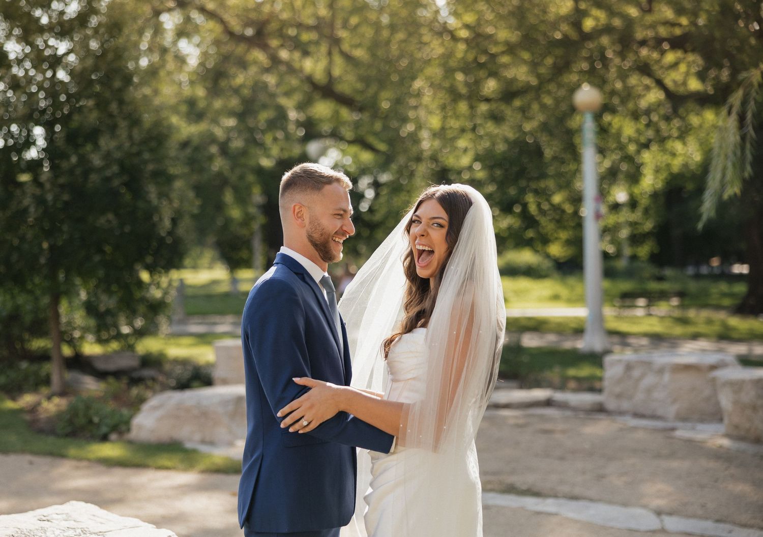 A couple in wedding attire share a romantic moment in a sunlit outdoor garden setting.
