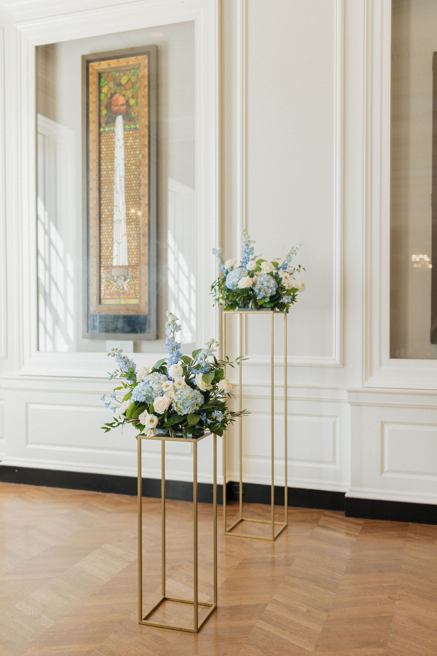 Modern gold geometric stands with white floral arrangements on herringbone floors in an elegant white room with framed art.