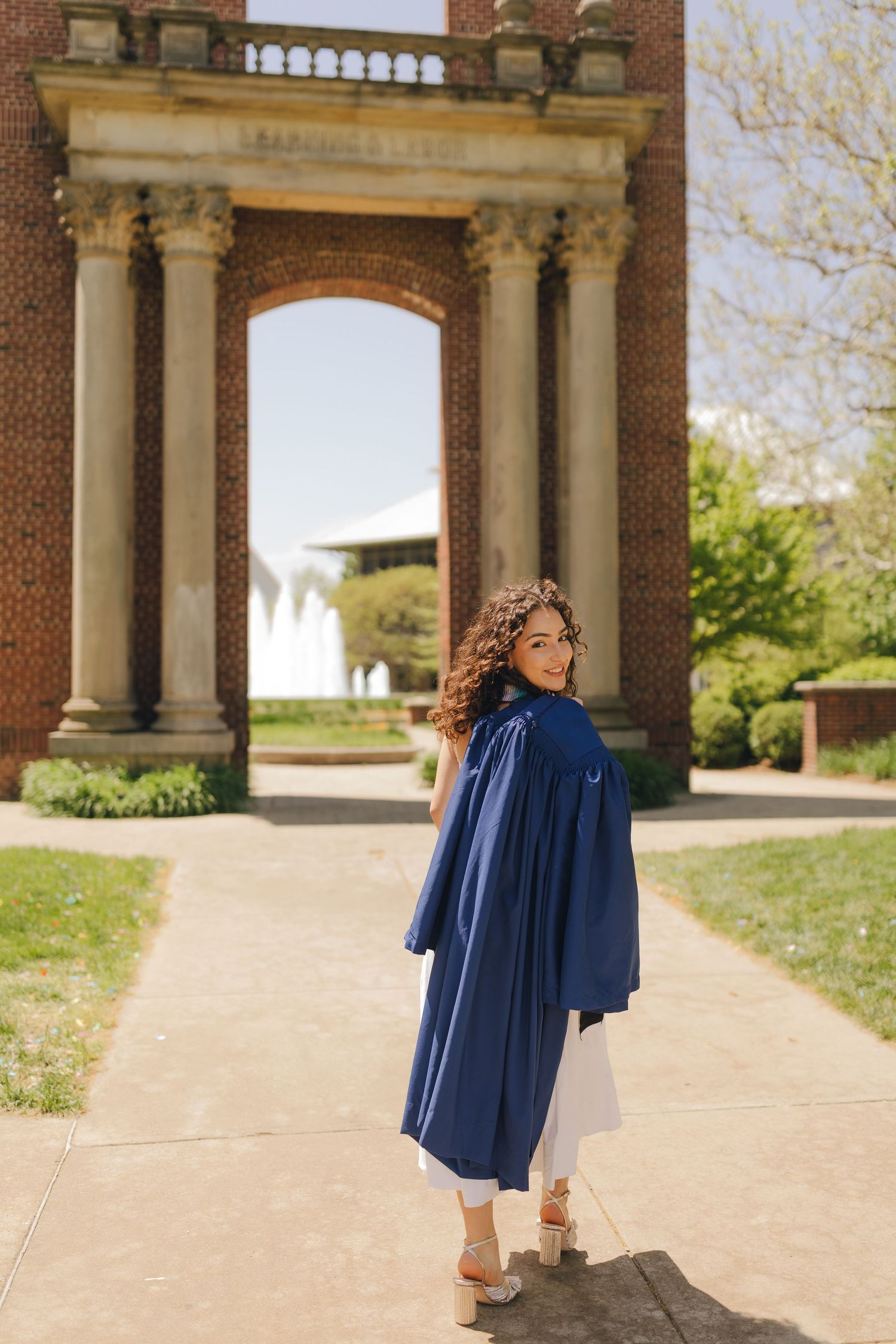 Graduate walking along a columned pathway on a sunny day wearing a navy blue graduation gown.