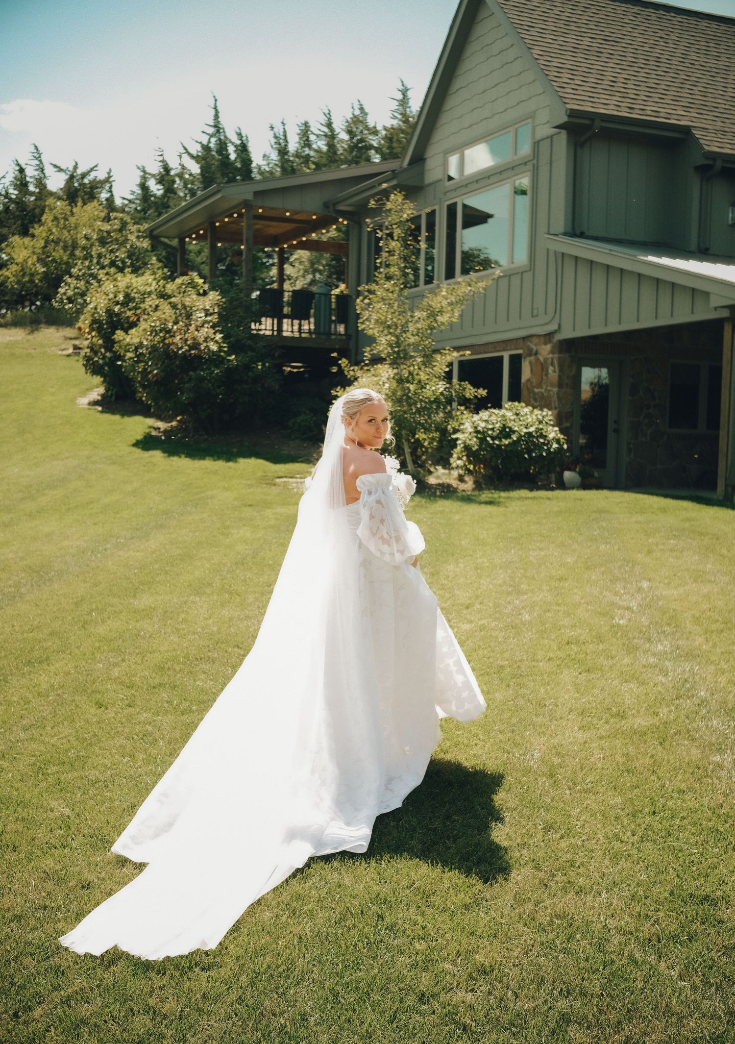 A flowing white wedding dress with a long train on a green lawn in front of a rustic house with a covered porch.