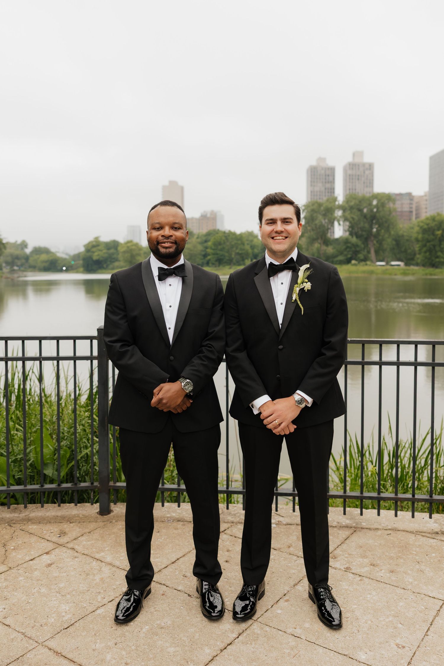 Two grooms in black tuxedos with boutonnieres pose by a lakeside railing in Central Park on their wedding day.