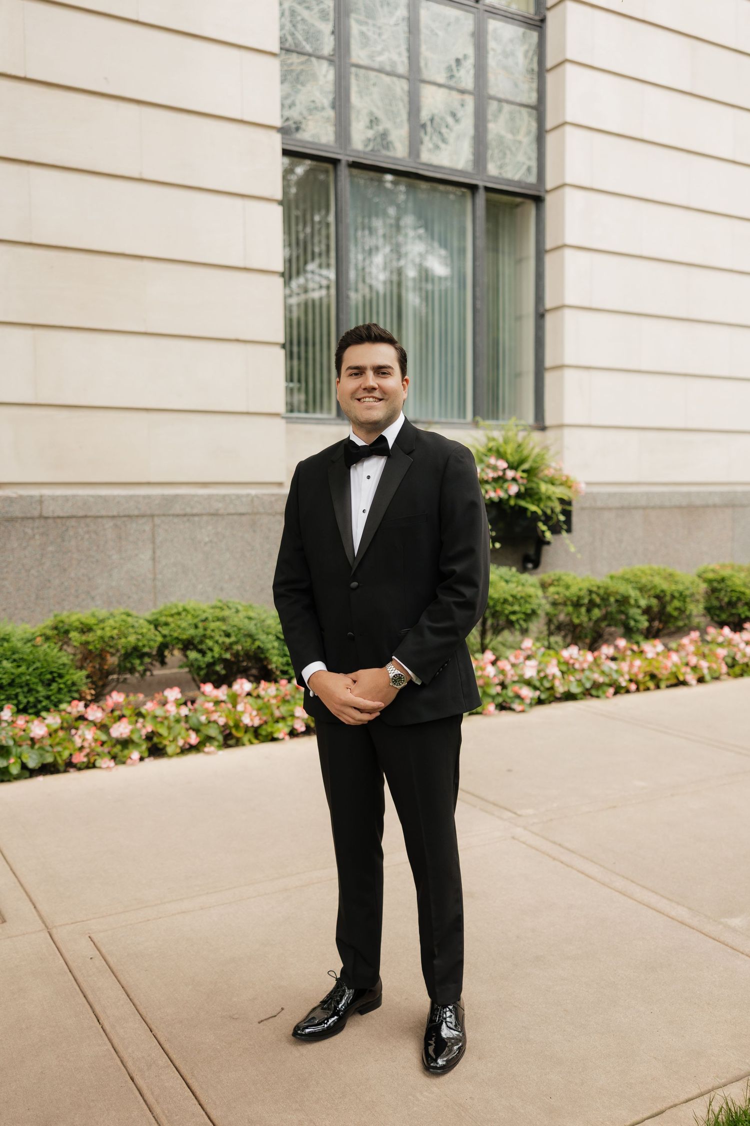 A smiling groom in a black tuxedo with bow tie stands on a sidewalk outside a stone building with pink flowers.