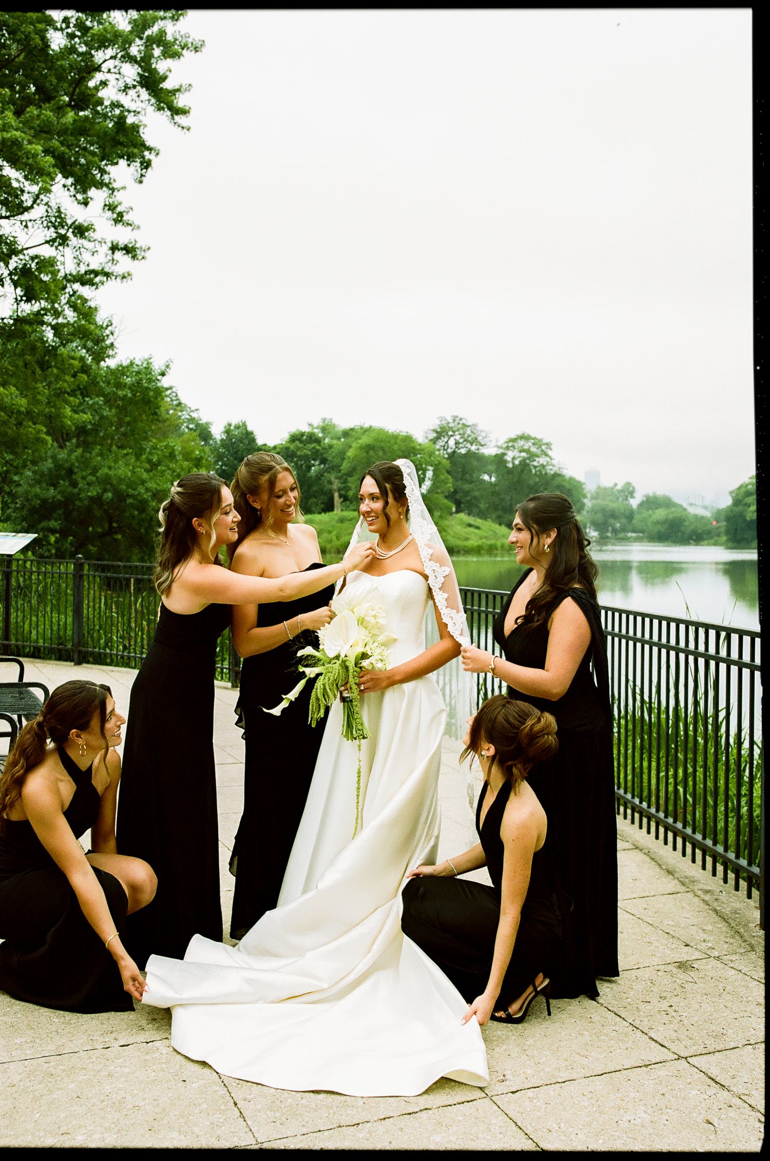 Bridesmaids in black dresses help bride with white gown and bouquet near a scenic park lake.