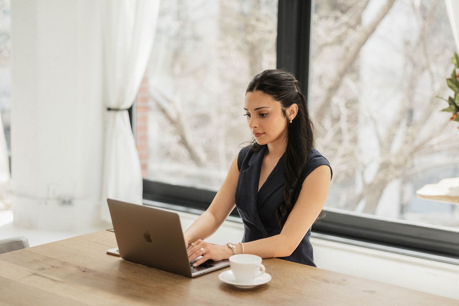 Young businesswoman working on laptop at wooden desk with coffee cup near bright windows.
