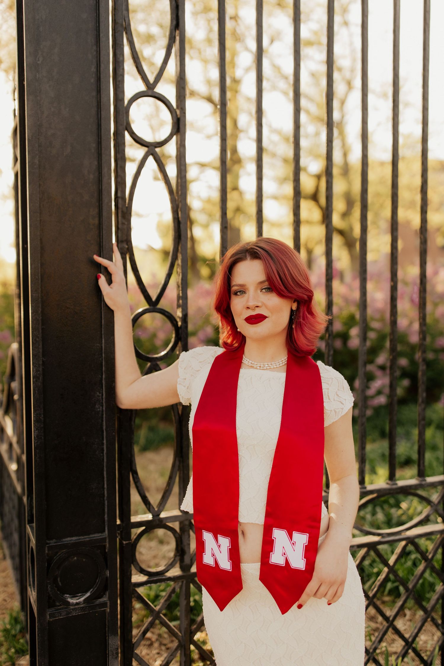Person in white dress and red scarf poses against ornate iron gate during golden hour sunset.