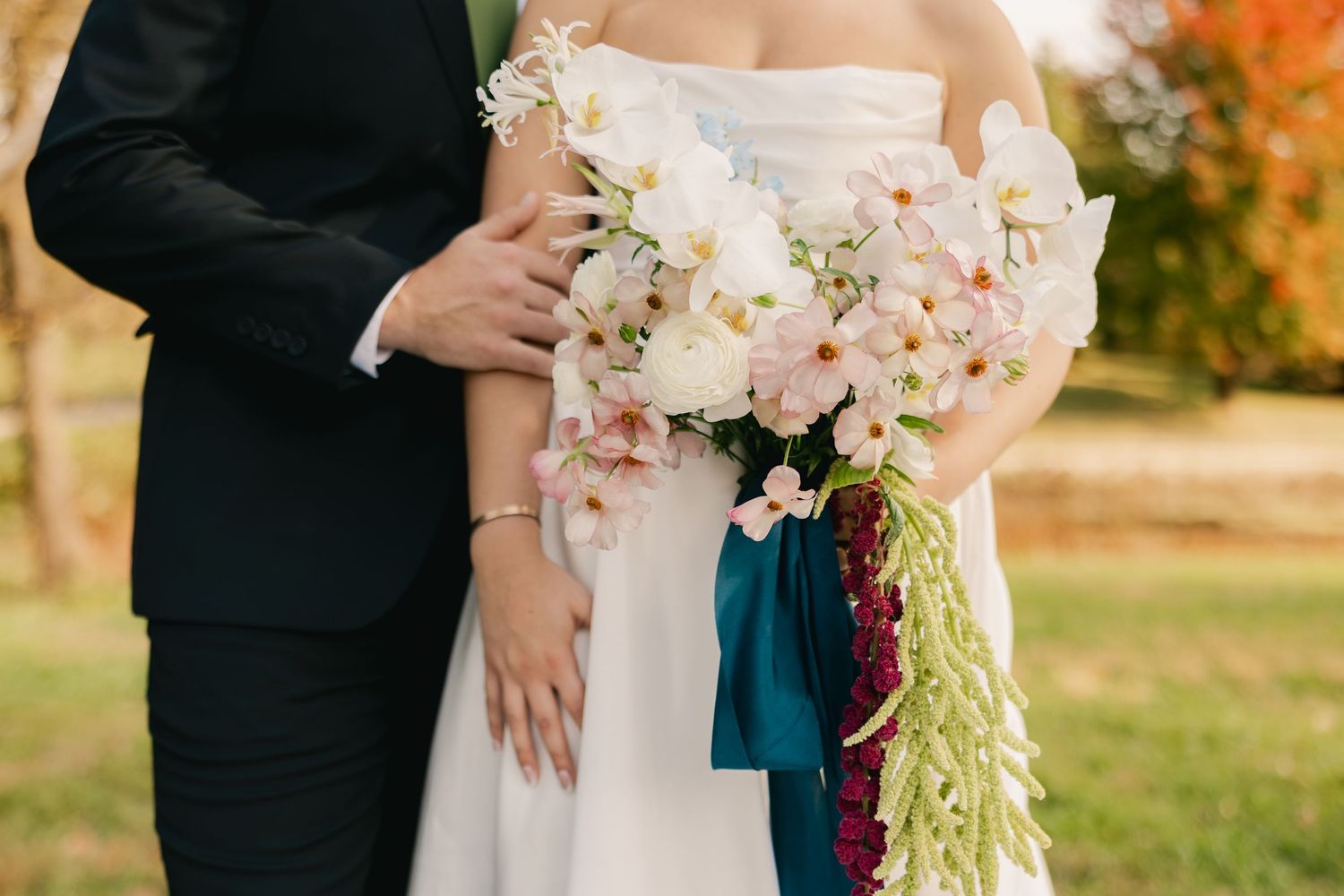 Bride in a white gown holds a cascading bouquet of pink and white flowers with teal ribbon beside a groom in a dark suit outdoors.