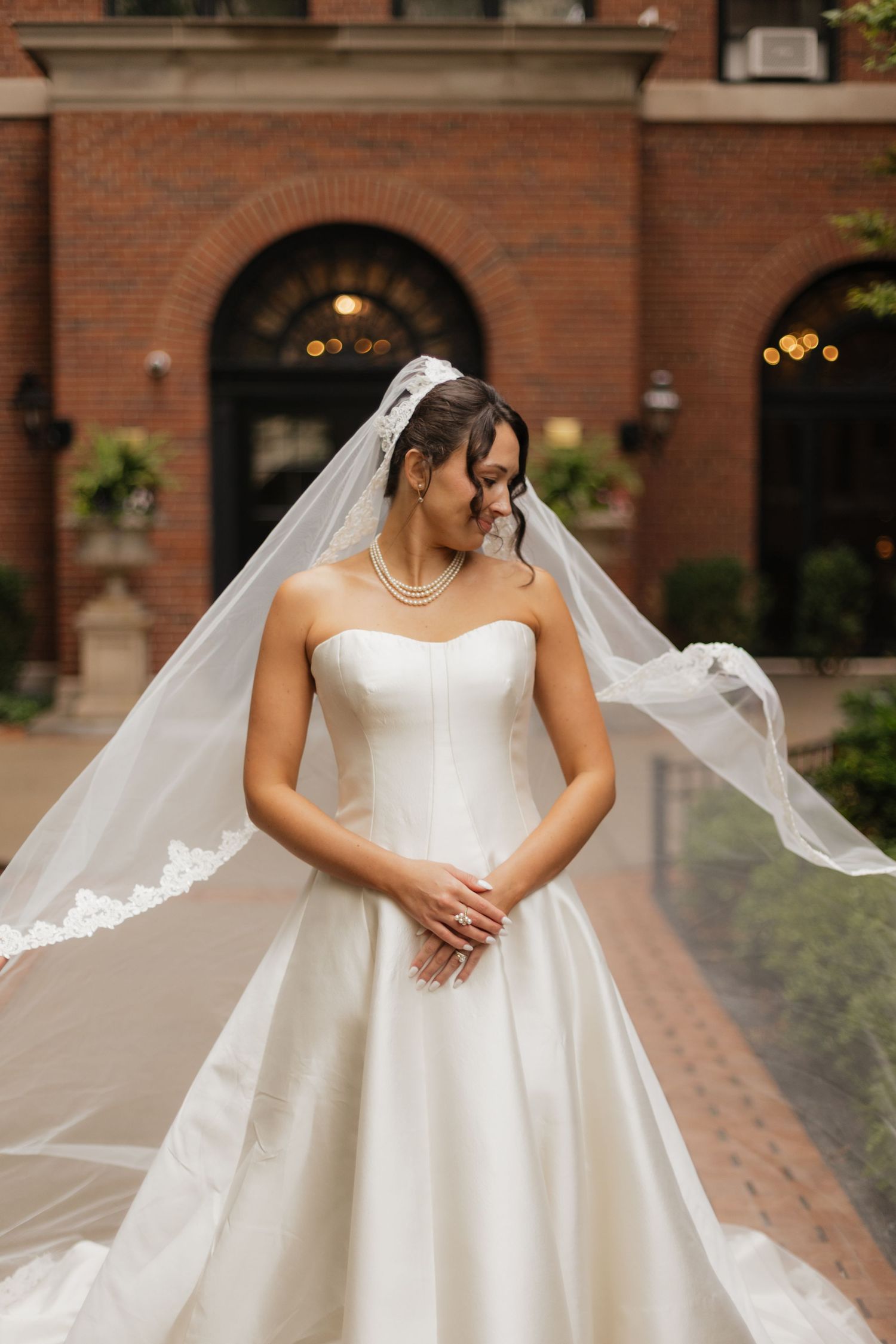 Bride in a strapless satin ballgown and lace-trimmed cathedral veil blowing in the wind, standing before a brick building with arched doors.