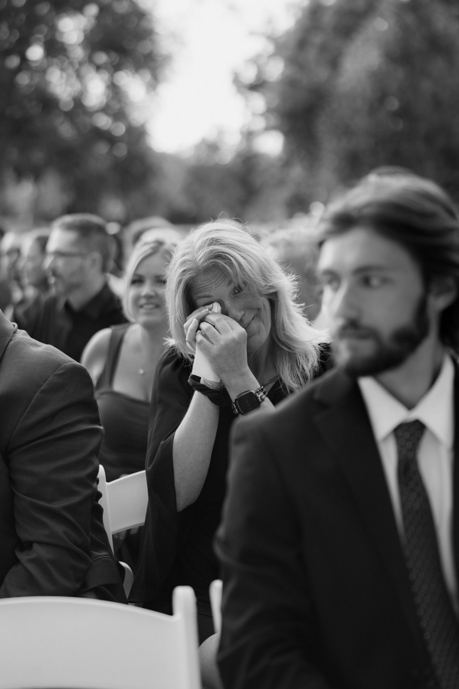 Black and white photo of an emotional woman wiping her tears at an outdoor wedding ceremony as guests sit nearby.