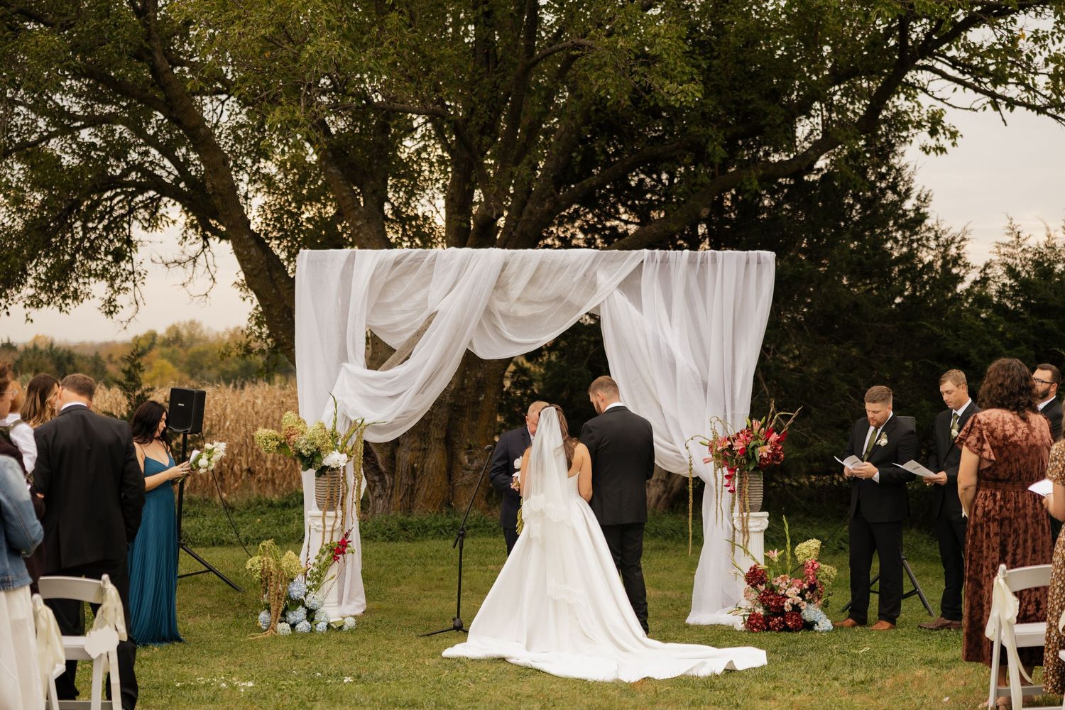 Bride in a long white gown and groom stand under a draped fabric canopy during an outdoor ceremony as guests watch beneath large trees.