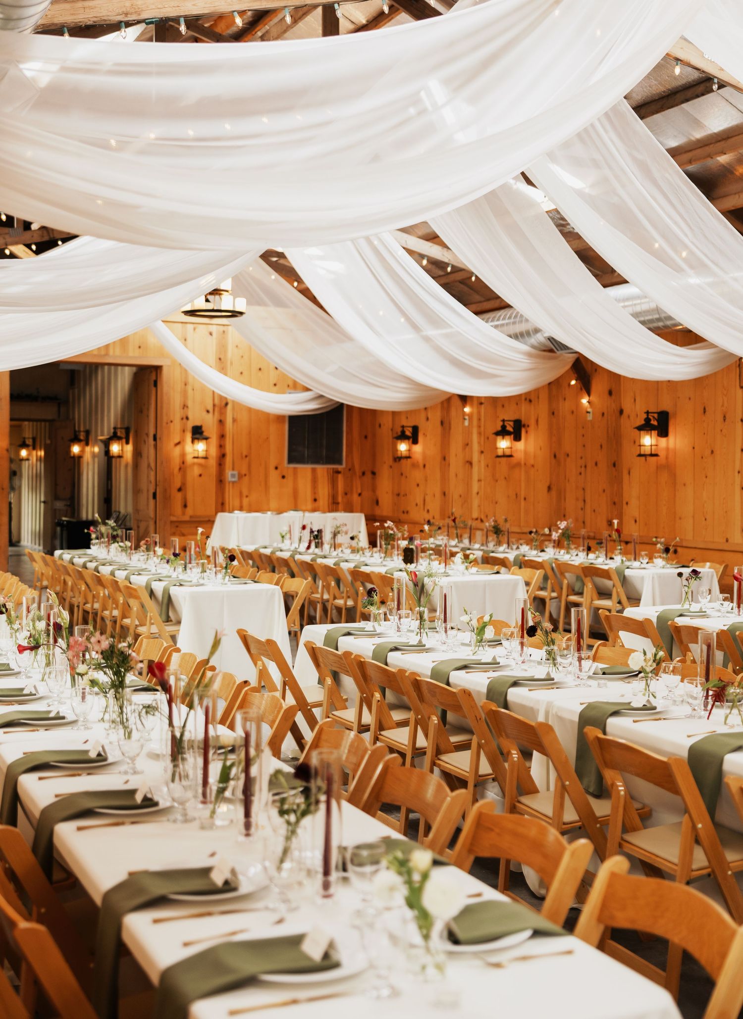 Rustic barn wedding reception with long tables, olive green napkins, wooden chairs, and white draped fabric across the wood-beamed ceiling.