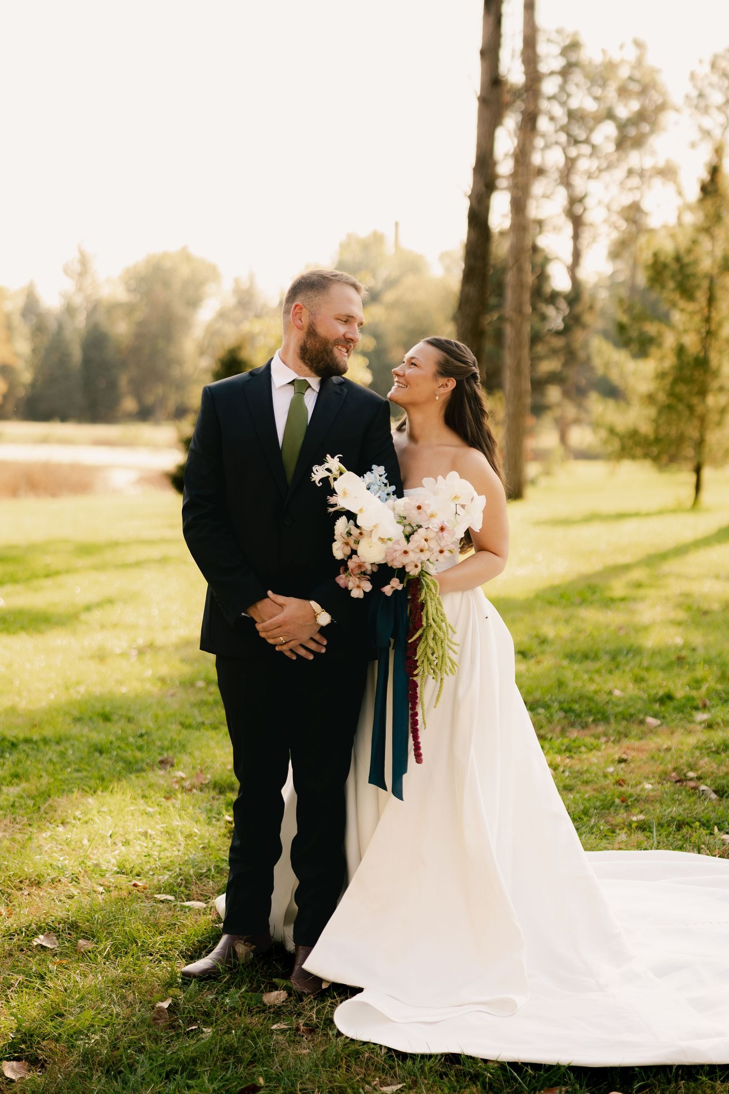 Bride in a white gown with a long train and groom in a dark suit smile at each other, holding a bouquet, on a sunny grassy lawn.