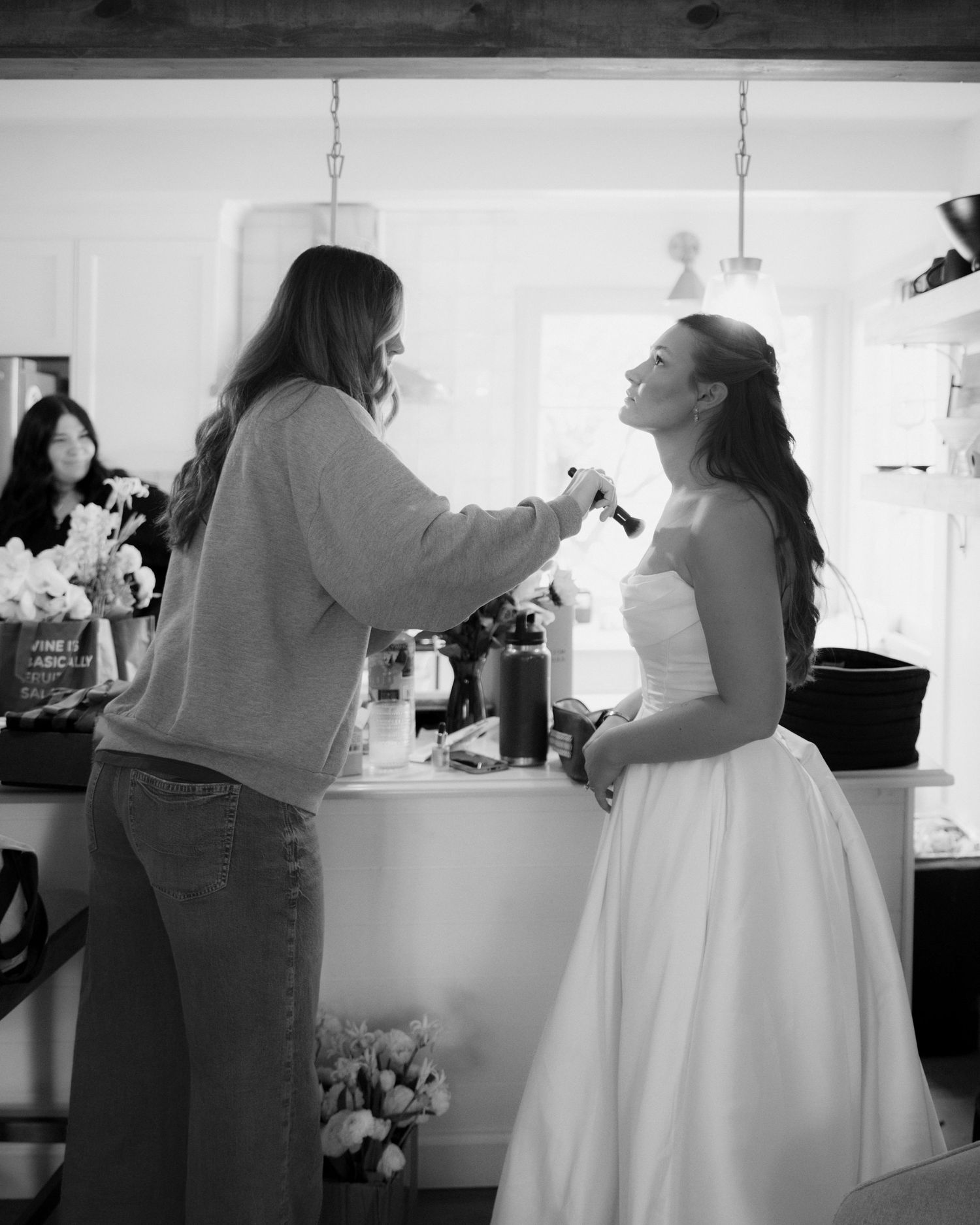 Makeup artist applies blush to a bride in a strapless wedding gown beside a counter with fresh flowers in a bright room, black and white.