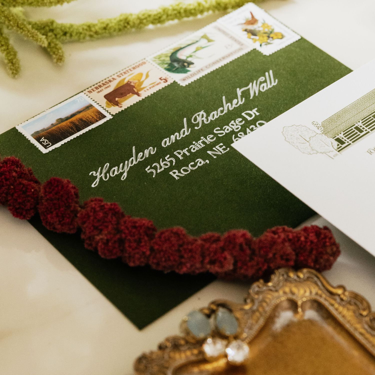 Dark green wedding invitation envelope with white calligraphy, vintage postage stamps, and burgundy ruffled trim beside a gold ornate frame.