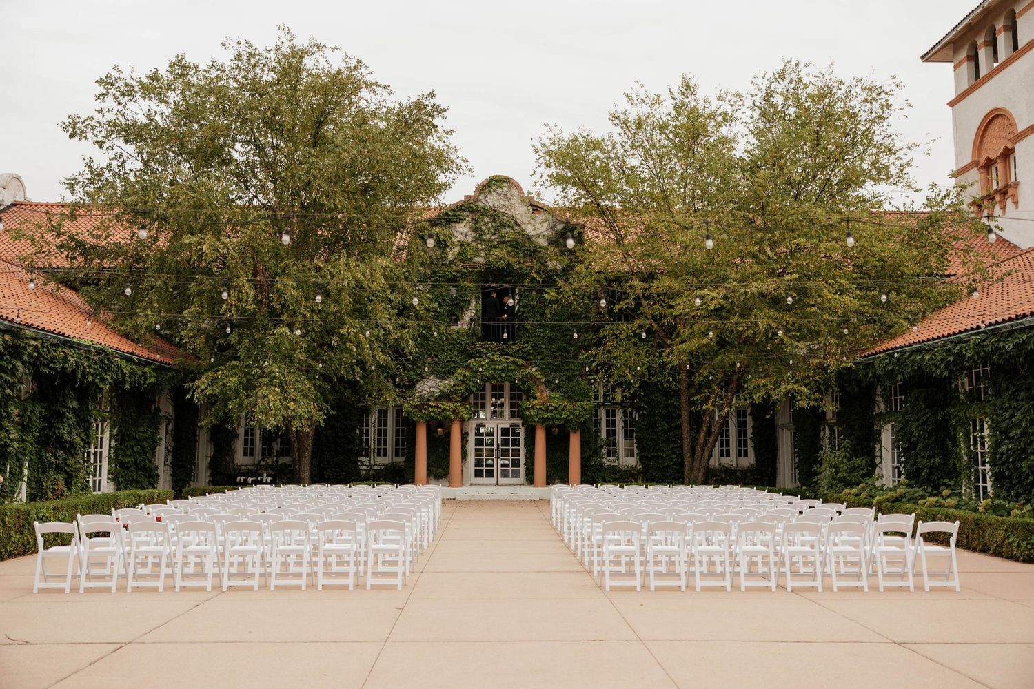 Rows of white chairs face an aisle in an outdoor wedding ceremony setup at an ivy-covered courtyard venue with tall shade trees.
