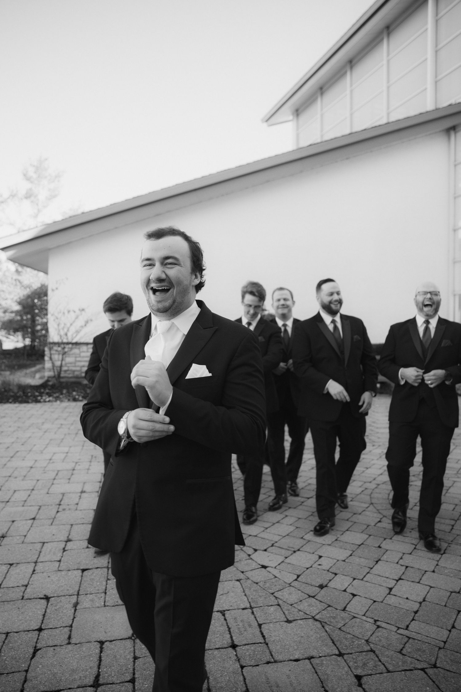 Black and white photo of a laughing groom in a suit walking with his groomsmen across a brick patio outside a white building.