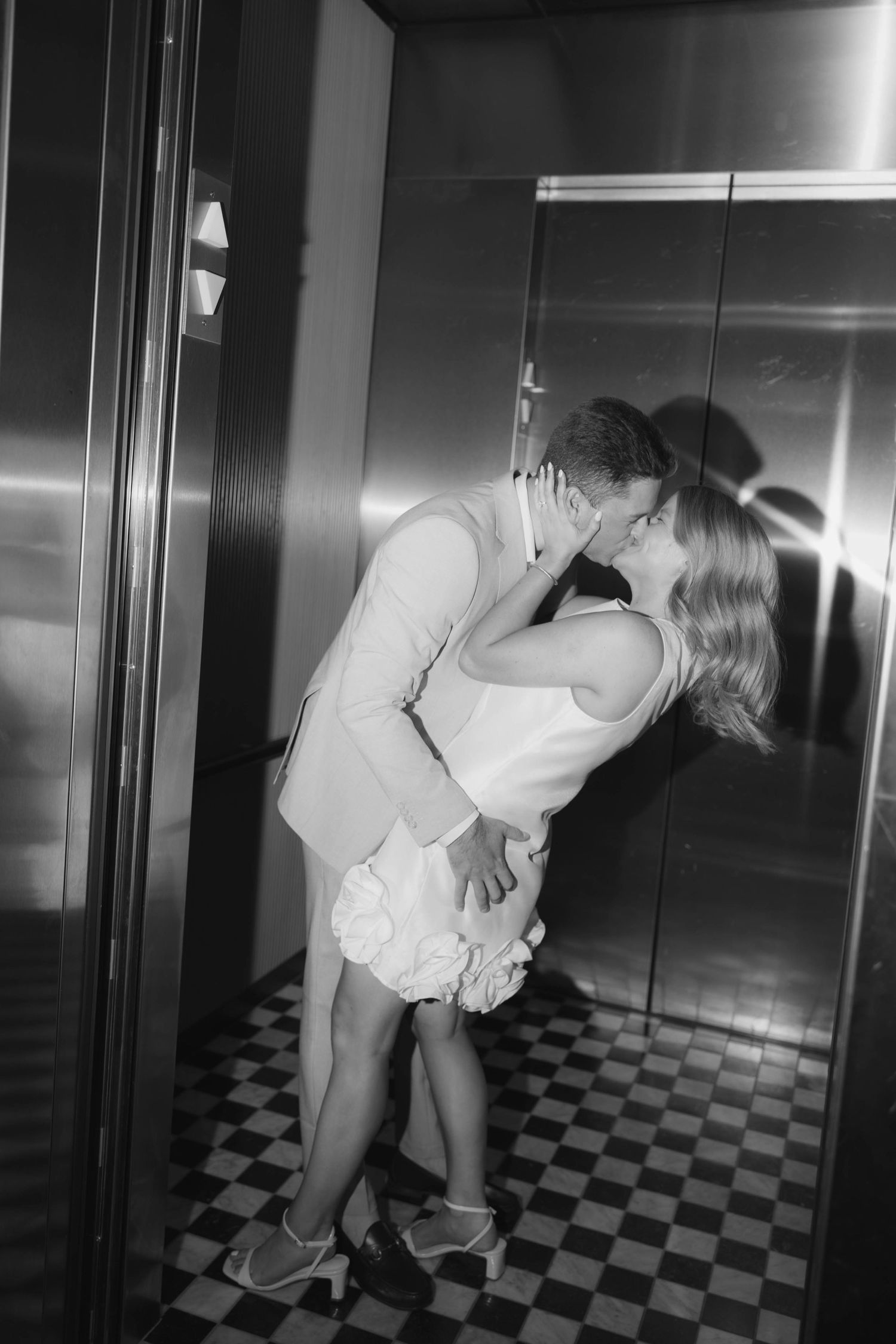 Black and white romantic photo of couple kissing in elevator with reflective metallic doors.