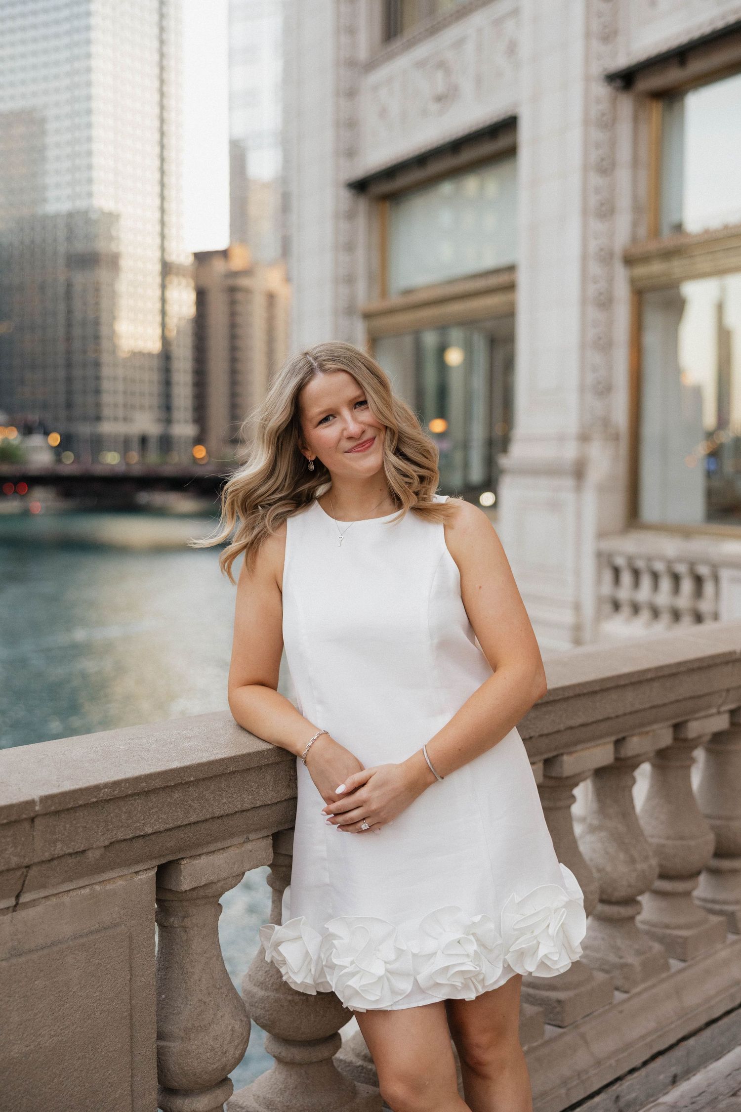 Woman in white ruffled mini dress posing by stone balustrade with Chicago River skyline backdrop.