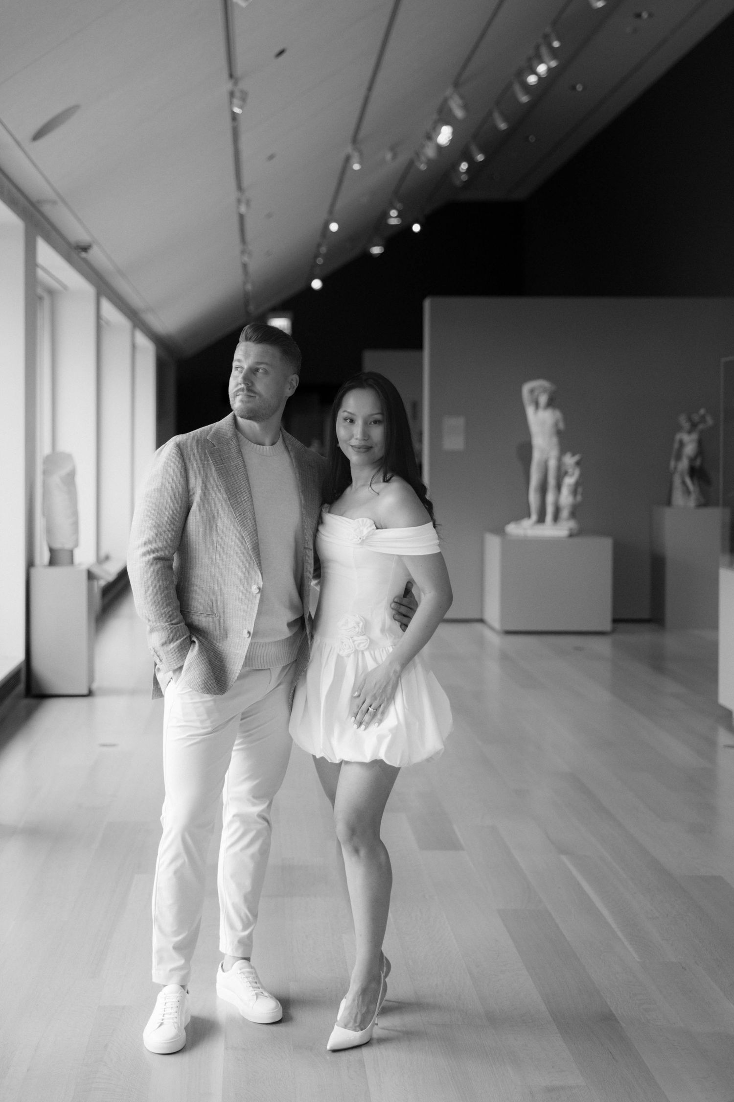 Black and white photo of a couple posing in an art museum gallery with classical sculptures in the background.