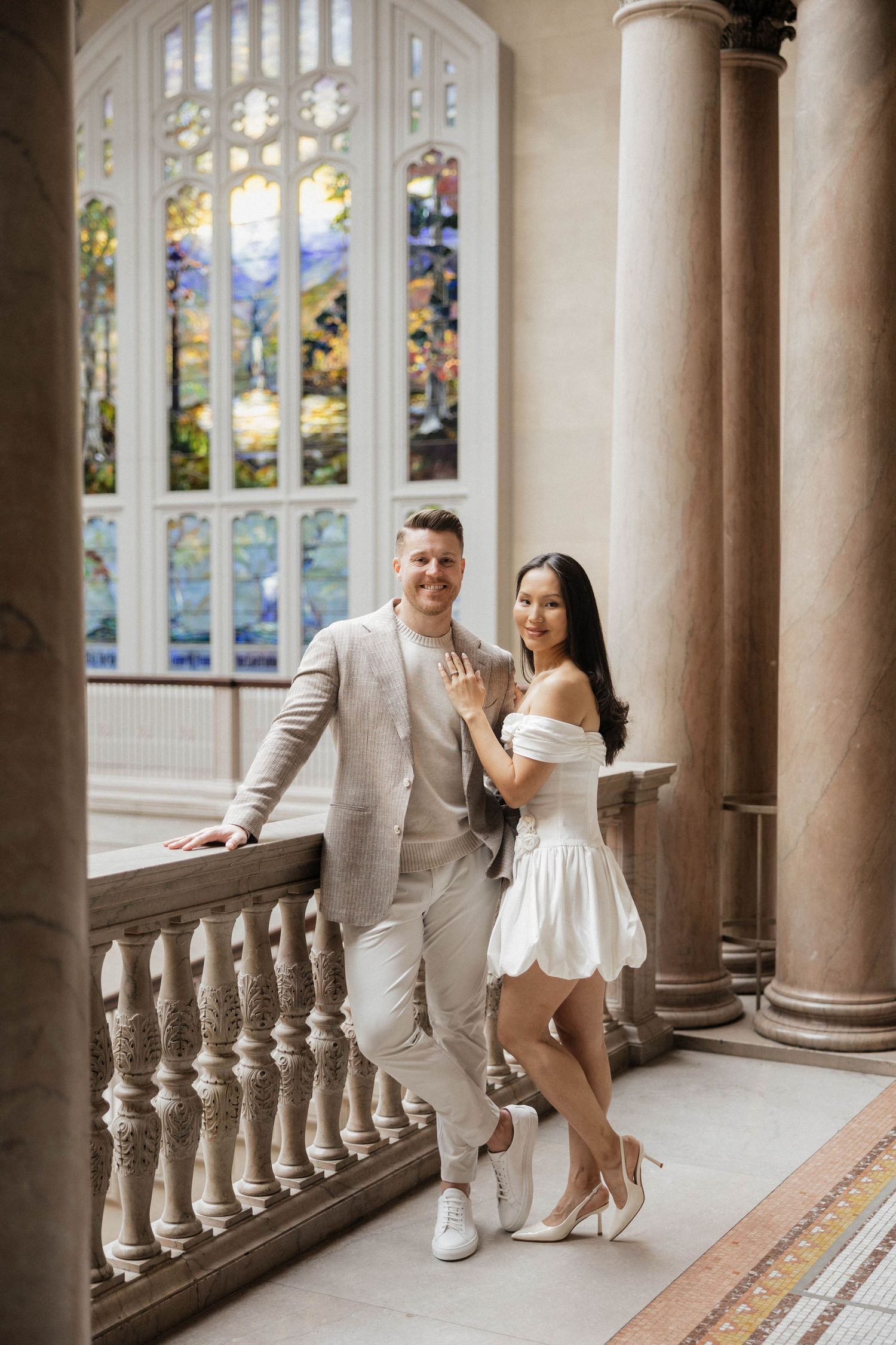 Couple posing in elegant hall with stained glass windows, woman showing engagement ring, wearing white dress and heels.