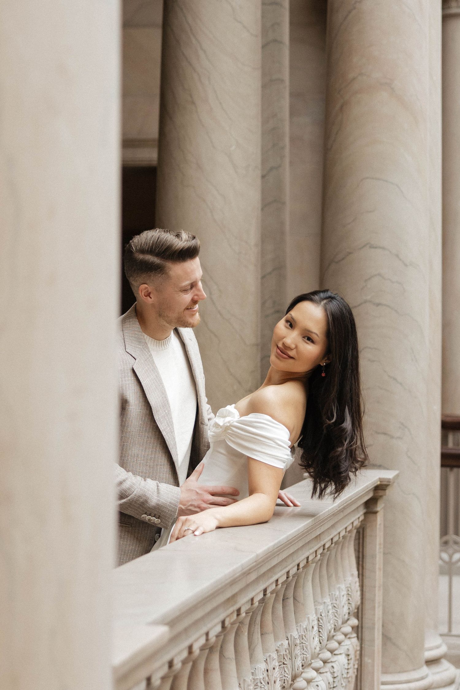Elegant couple posing on a classical balcony with marble columns, woman in white off-shoulder dress smiling at camera.