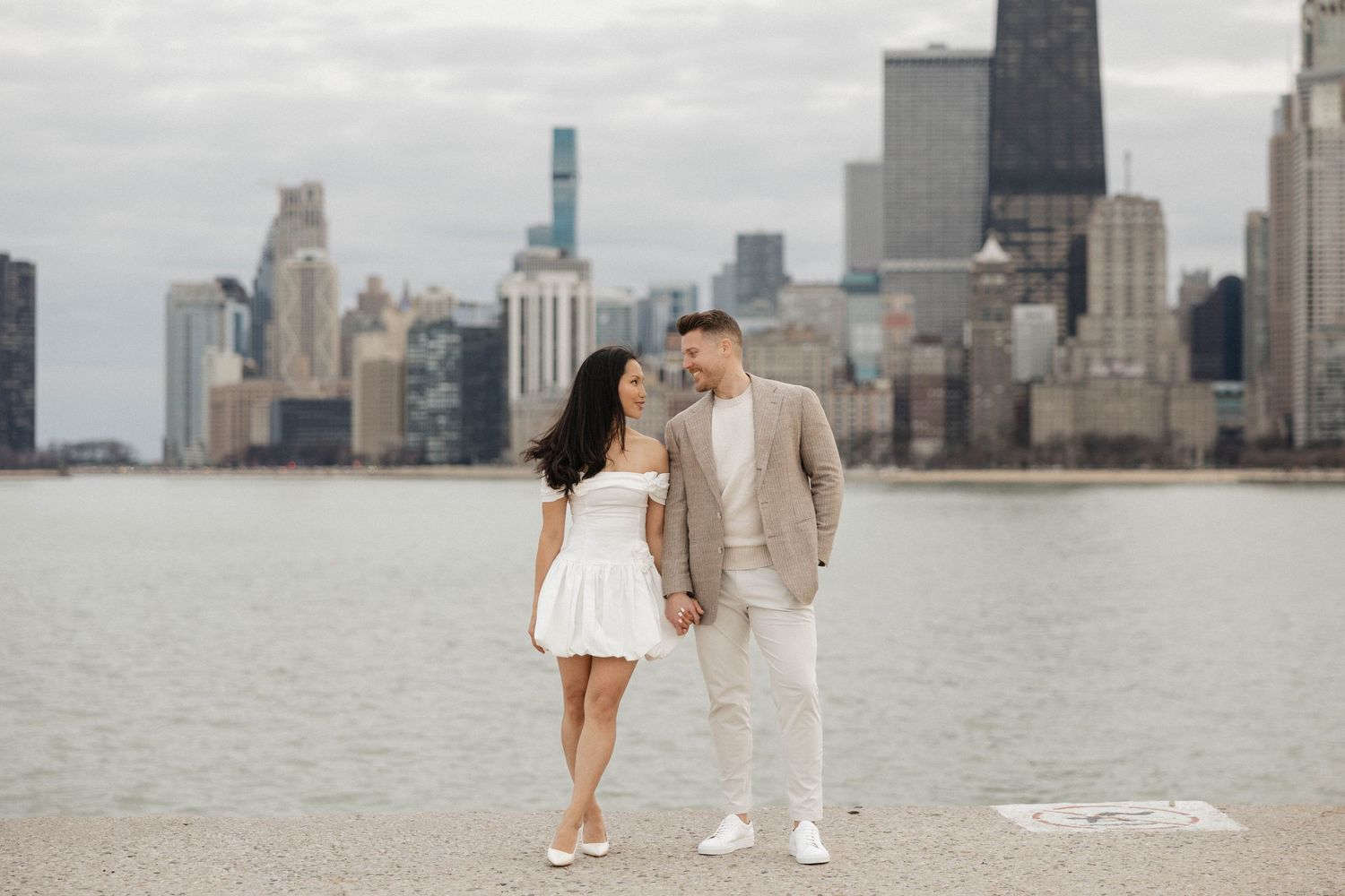 Engaged couple holding hands on Chicago waterfront, woman in white off-shoulder dress, man in beige suit, skyline behind.