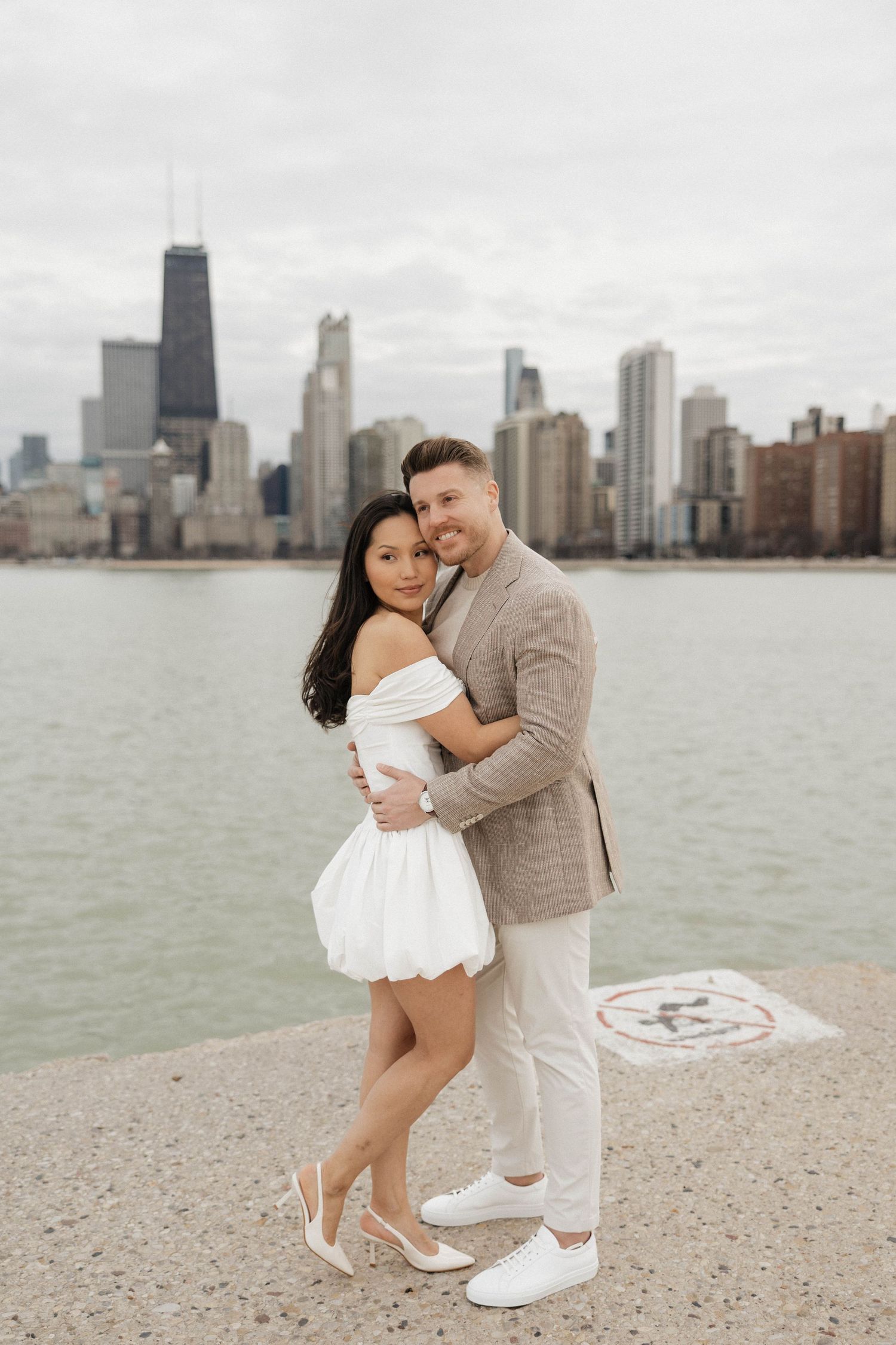 Couple embracing on Chicago lakefront pier with city skyline backdrop, woman in white mini dress, man in tan blazer.