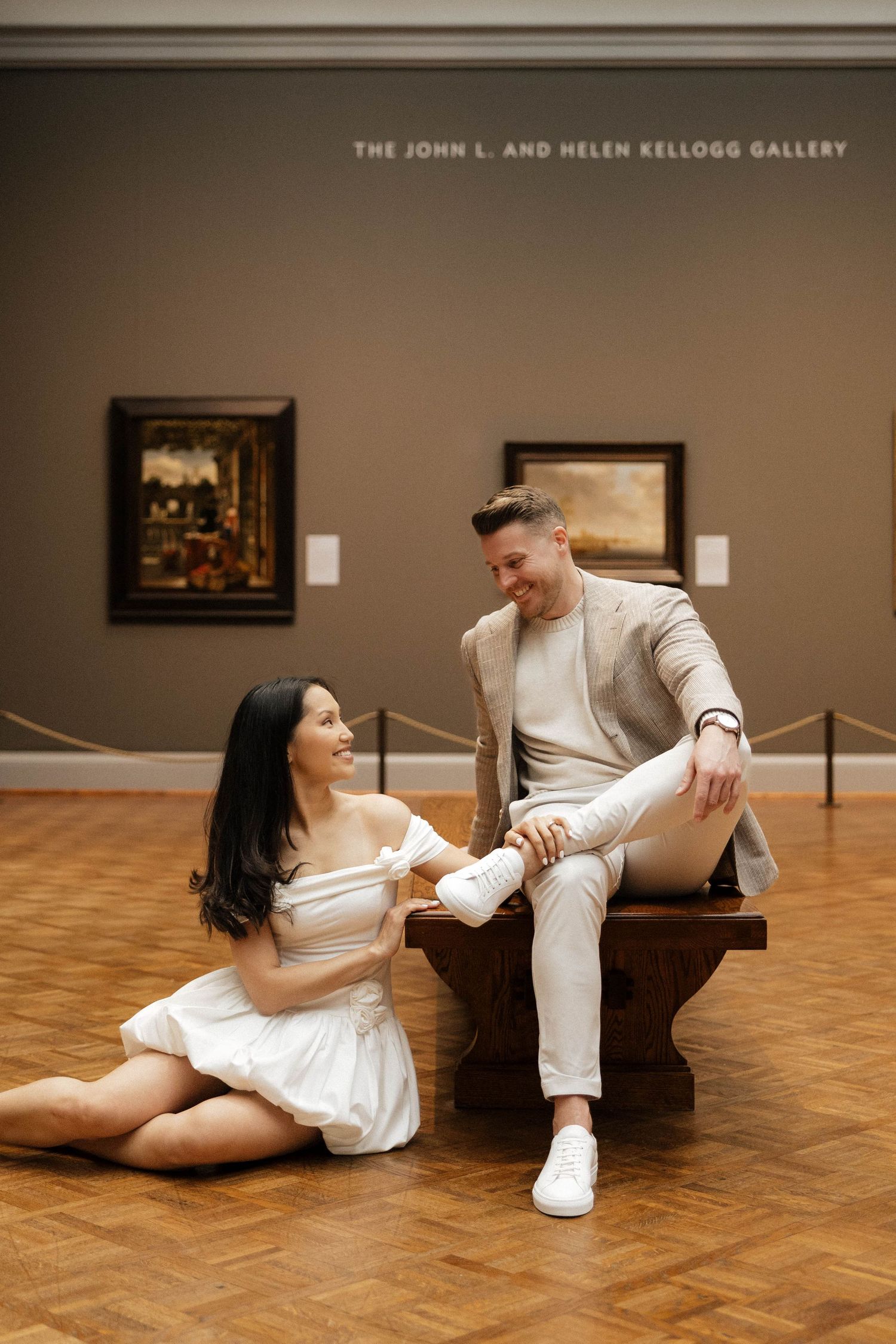 Couple posing in the John L. and Helen Kellogg Gallery, woman in white dress seated on floor, man in beige suit on bench.