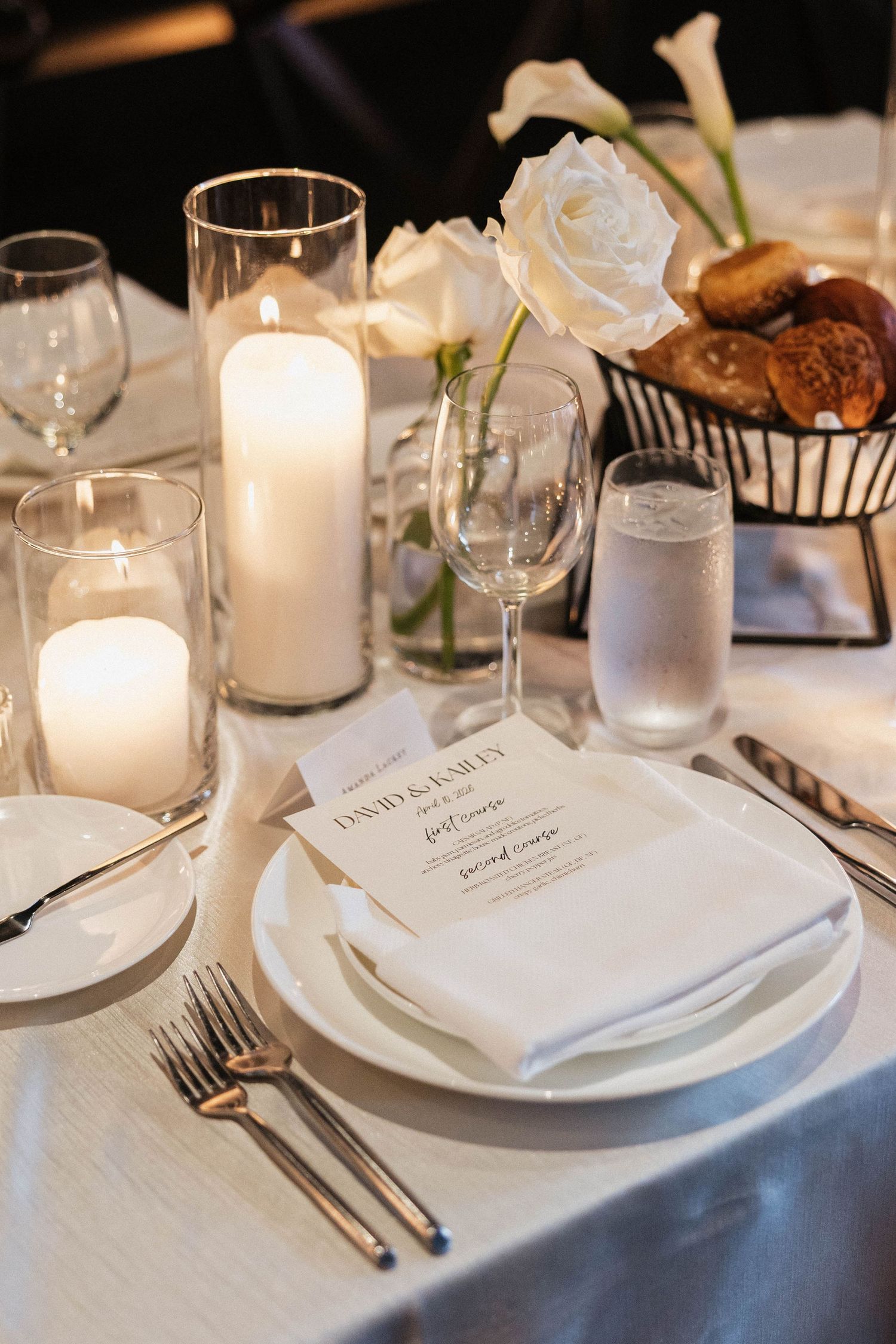 Elegant wedding table setting with white candles, roses, wine glasses, and a personalized menu card on white linens.