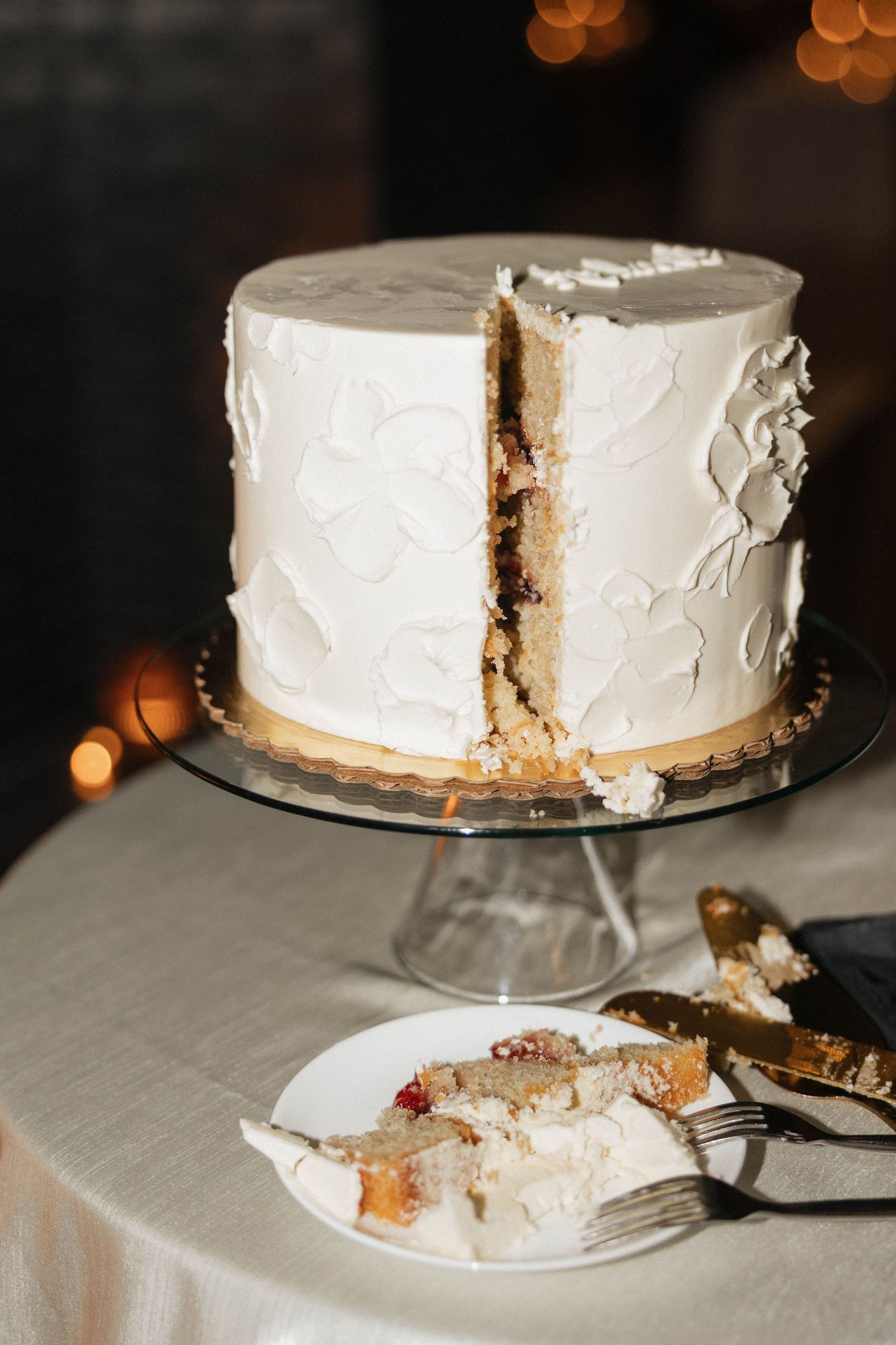 White wedding cake with textured frosting on glass stand, slice removed revealing layers, with plated piece and gold forks.