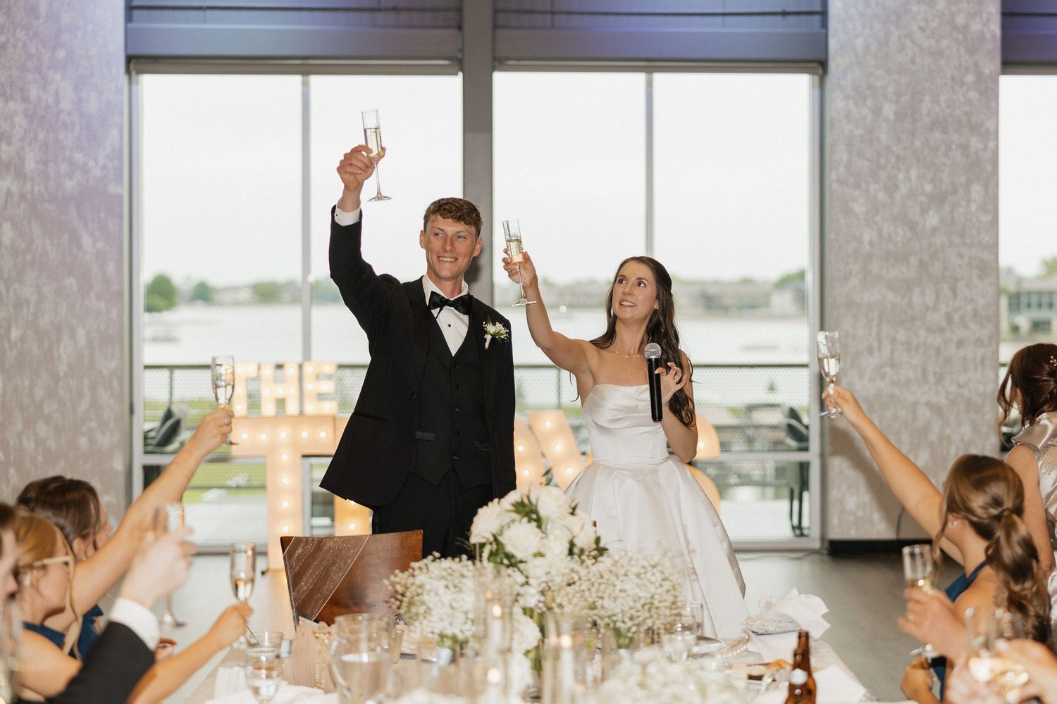Bride and groom raise champagne glasses during wedding reception toast with guests at waterfront venue.