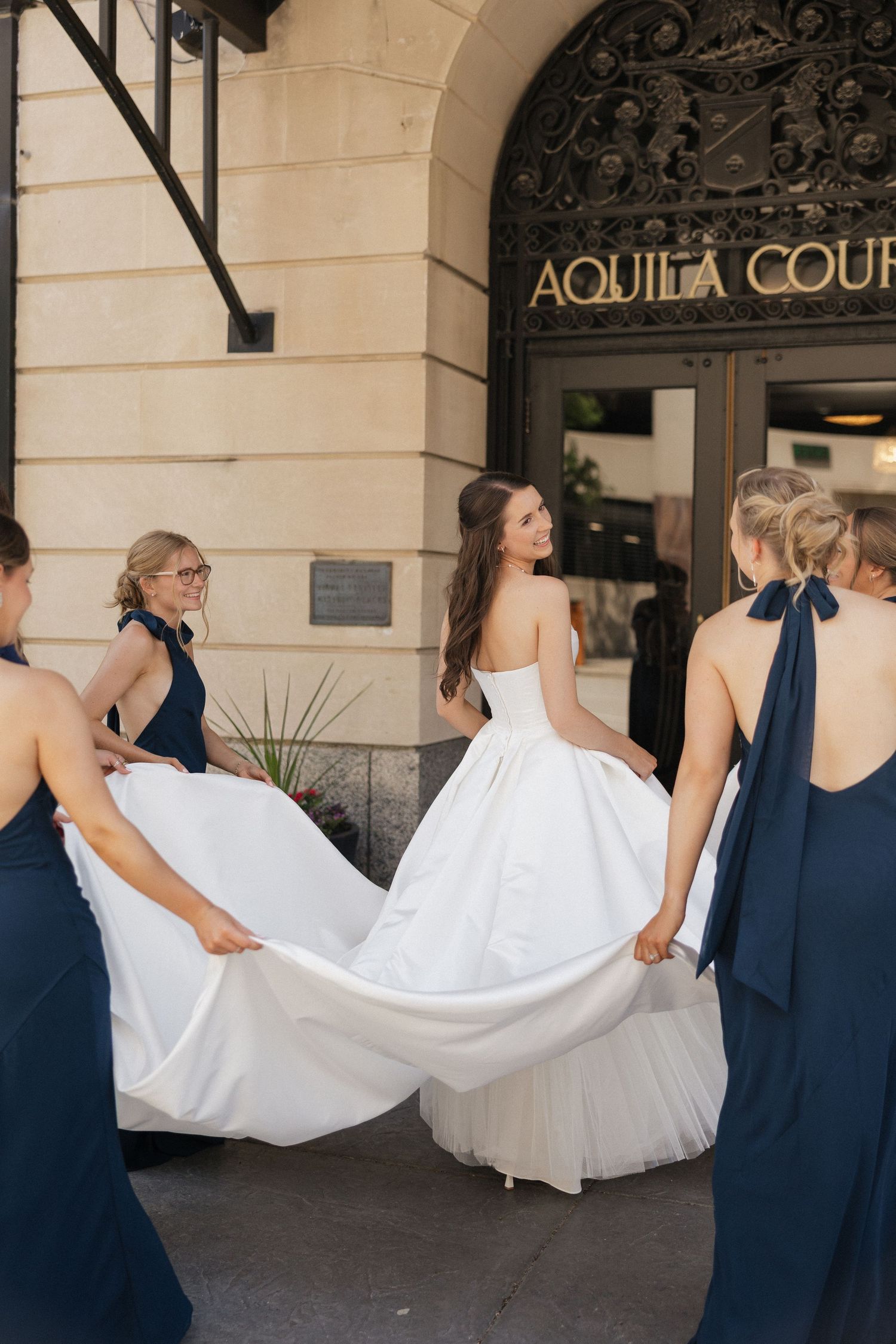 Bridesmaids in navy dresses hold the train of a white bridal gown outside Aquila Court venue.