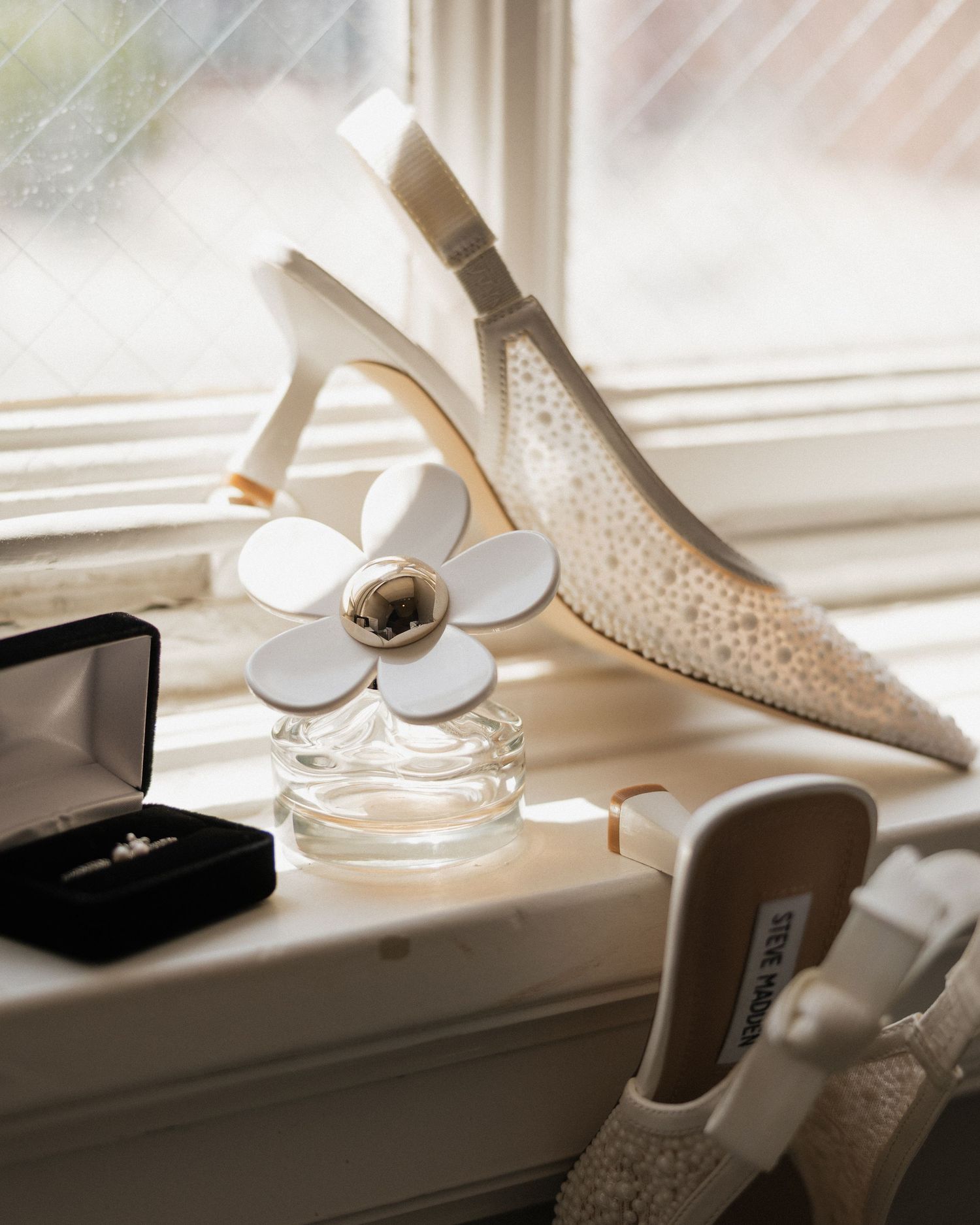 Elegant bridal accessories on a windowsill featuring ivory heels, a daisy perfume bottle, and a diamond ring in a black box.
