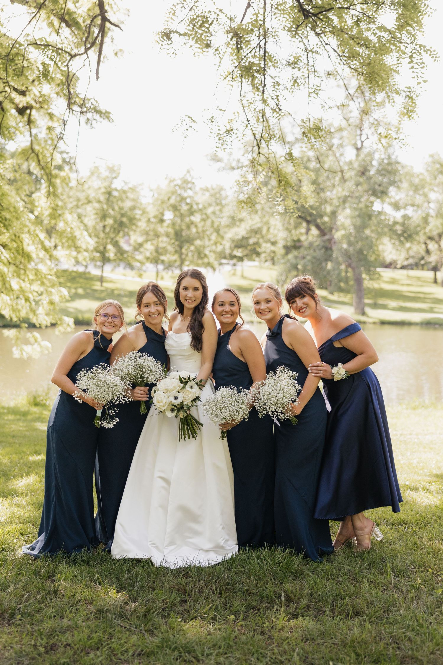 Bride in white gown poses with five bridesmaids in navy blue dresses holding white bouquets outdoors near a pond.