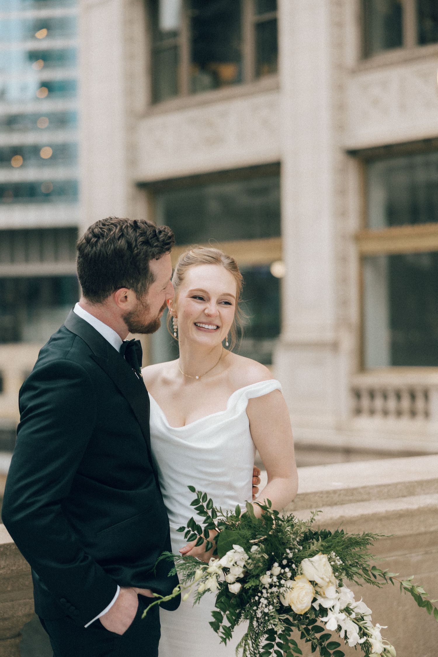A couple in wedding attire share an intimate moment outside a building with a romantic floral bouquet.