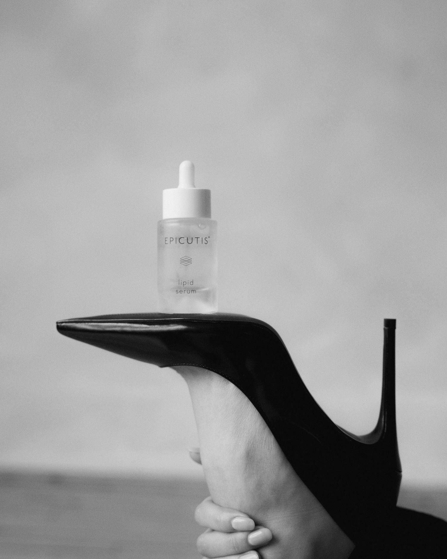 A black high heel shoe held sideways balancing a small white cosmetic dropper bottle in black and white photography.