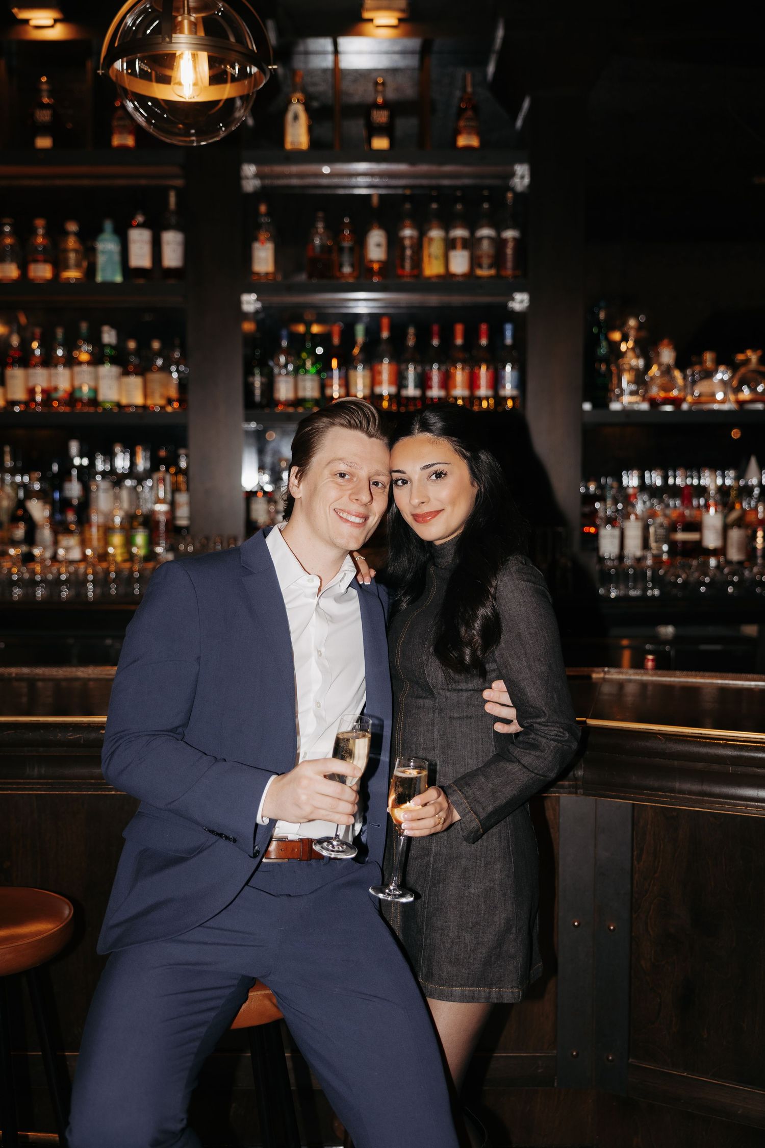 A couple shares drinks at a dimly lit bar with bottles displayed in the background.